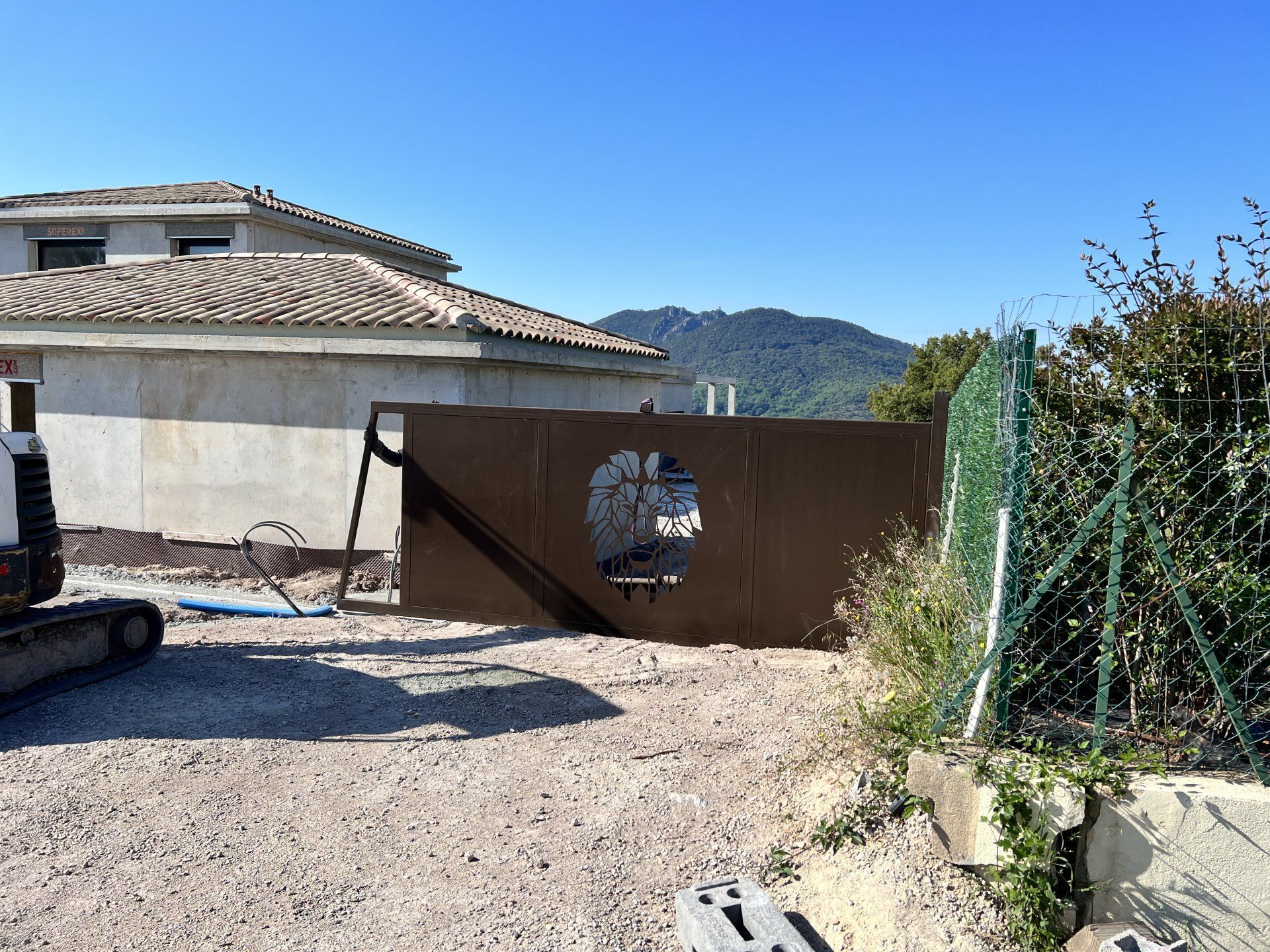 Portail en métal marron dans une allée de gravier à côté d'une maison, avec des montagnes et un ciel bleu clair en arrière-plan.
