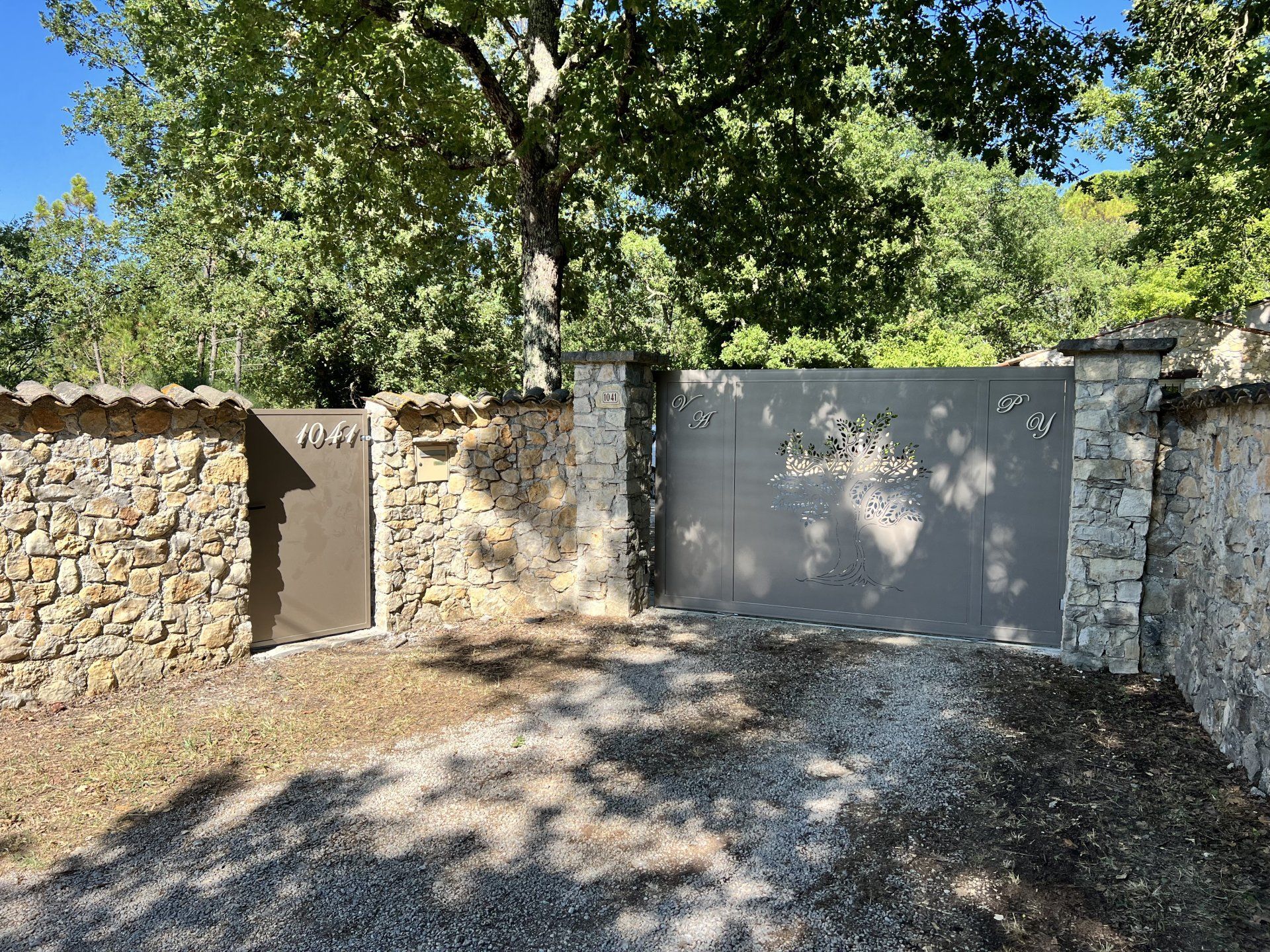 Mur de pierre et portail gris bordant une allée de gravier sous des arbres feuillus baignés de soleil