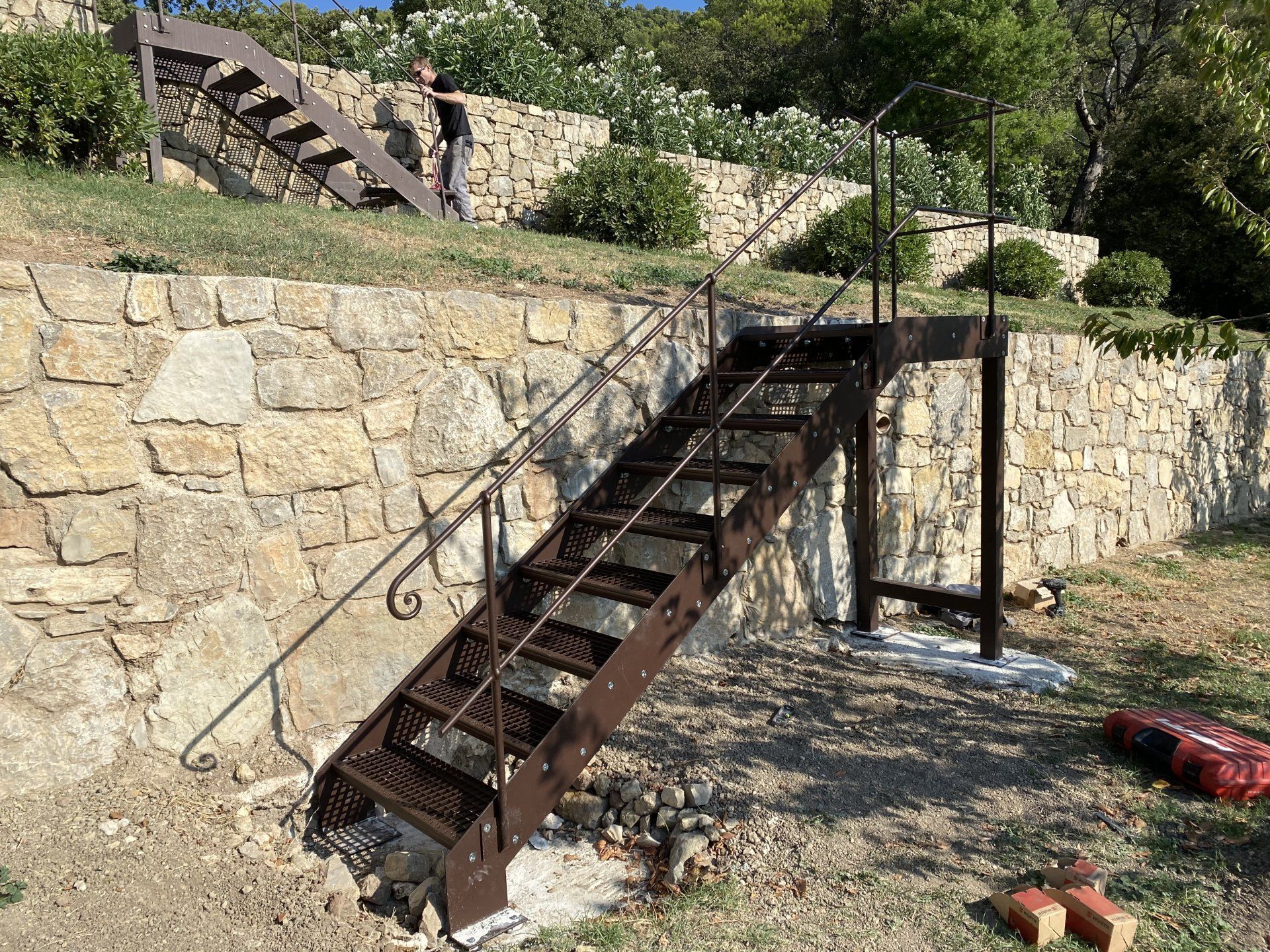 Escalier métallique rouillé fixé à un mur de soutènement en pierre dans une cour extérieure ensoleillée.