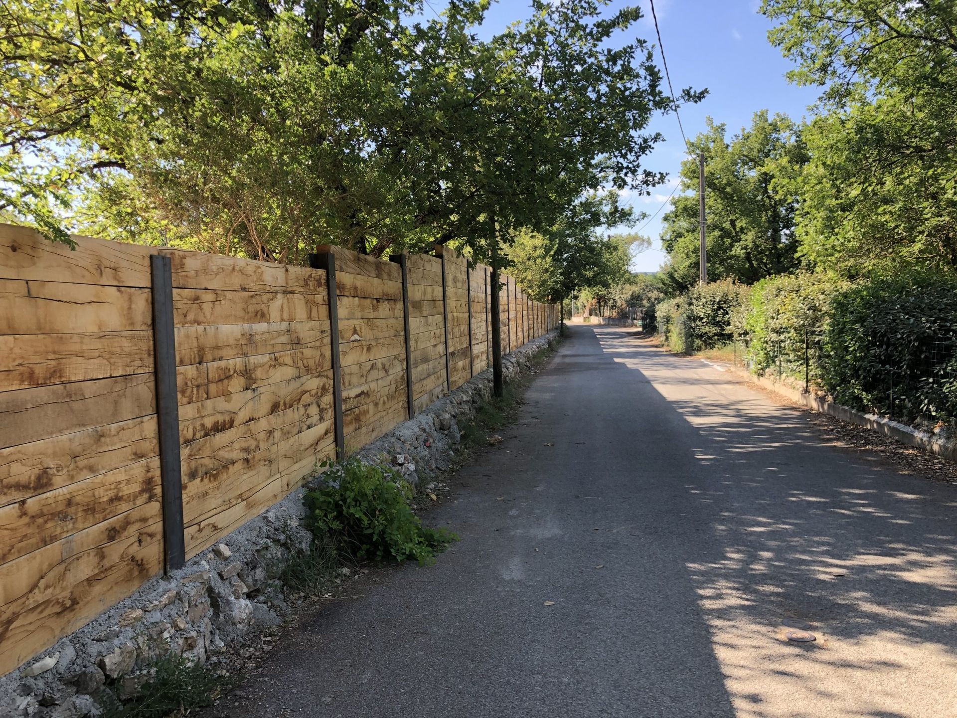 Chemin de terre ensoleillé longeant une clôture en bois et des arbres denses sous un ciel bleu limpide