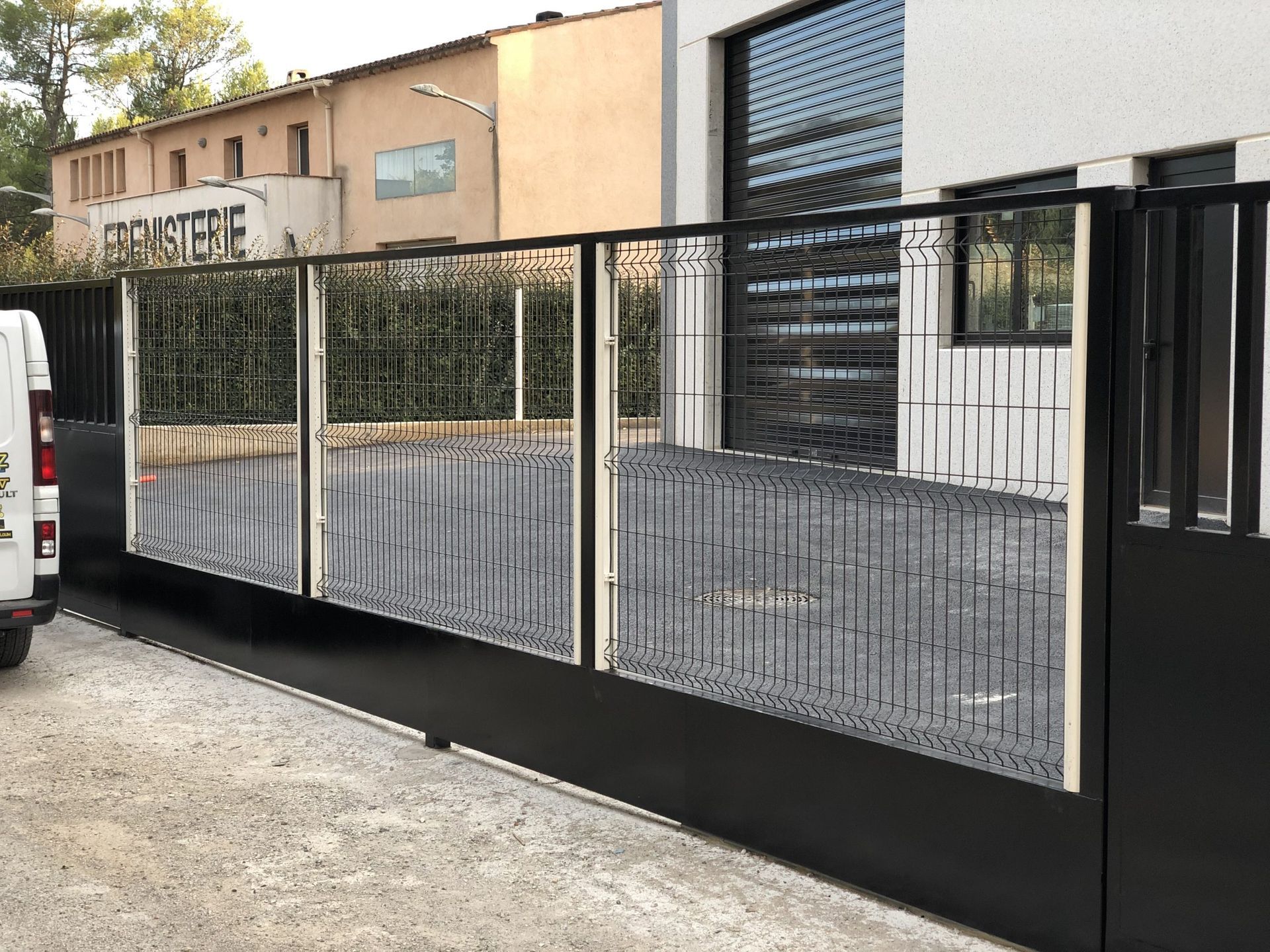 Portail coulissant en métal noir à barreaux verticaux devant un bâtiment industriel et une allée de gravier.