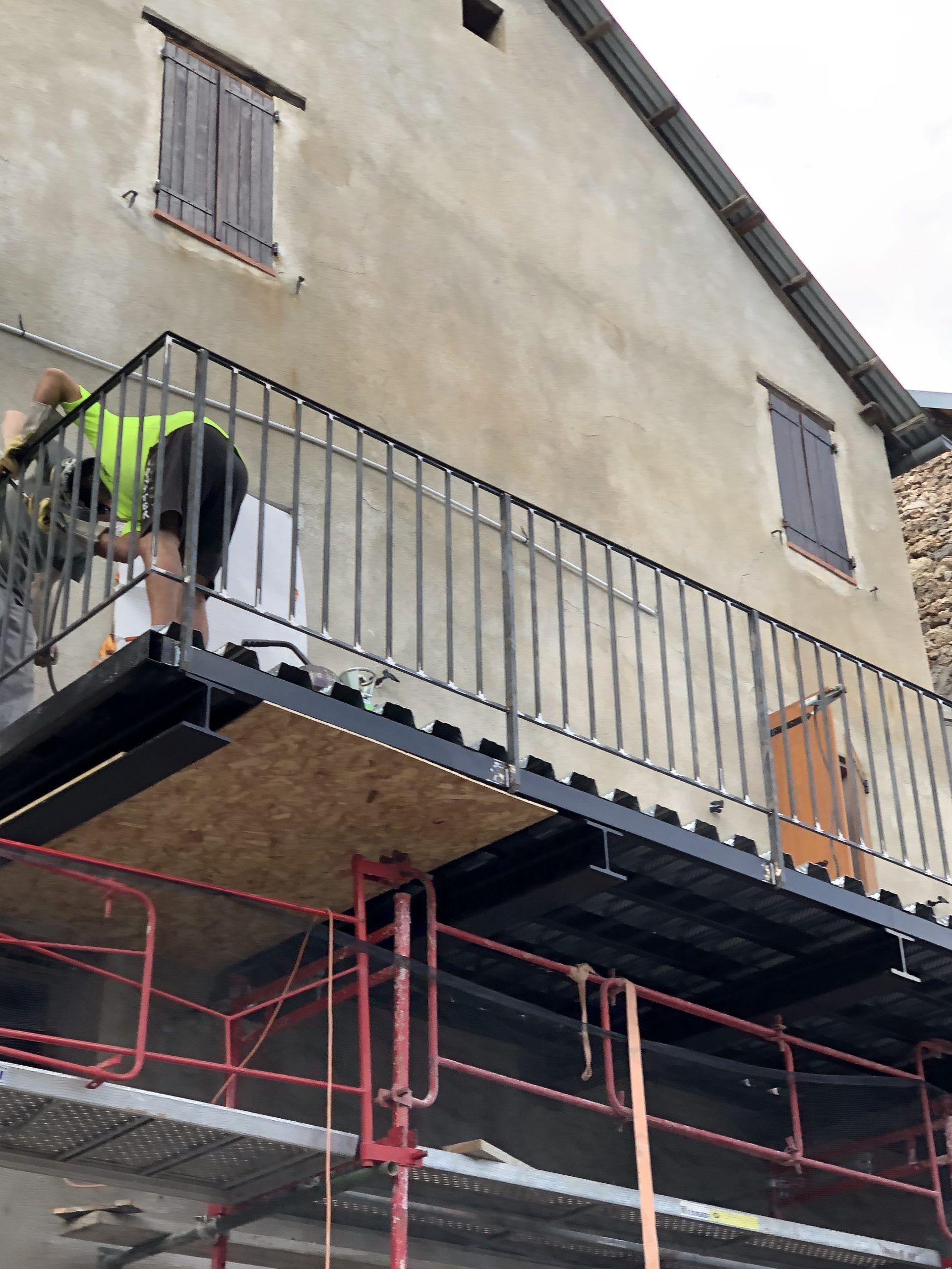 Un ouvrier peint la façade d'un immeuble depuis un échafaudage, sous un balcon.