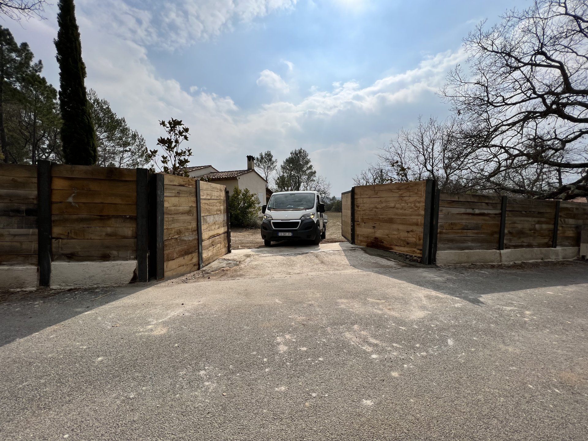 Un SUV noir franchit un portail en bois sur une allée de gravier sous un ciel partiellement nuageux.
