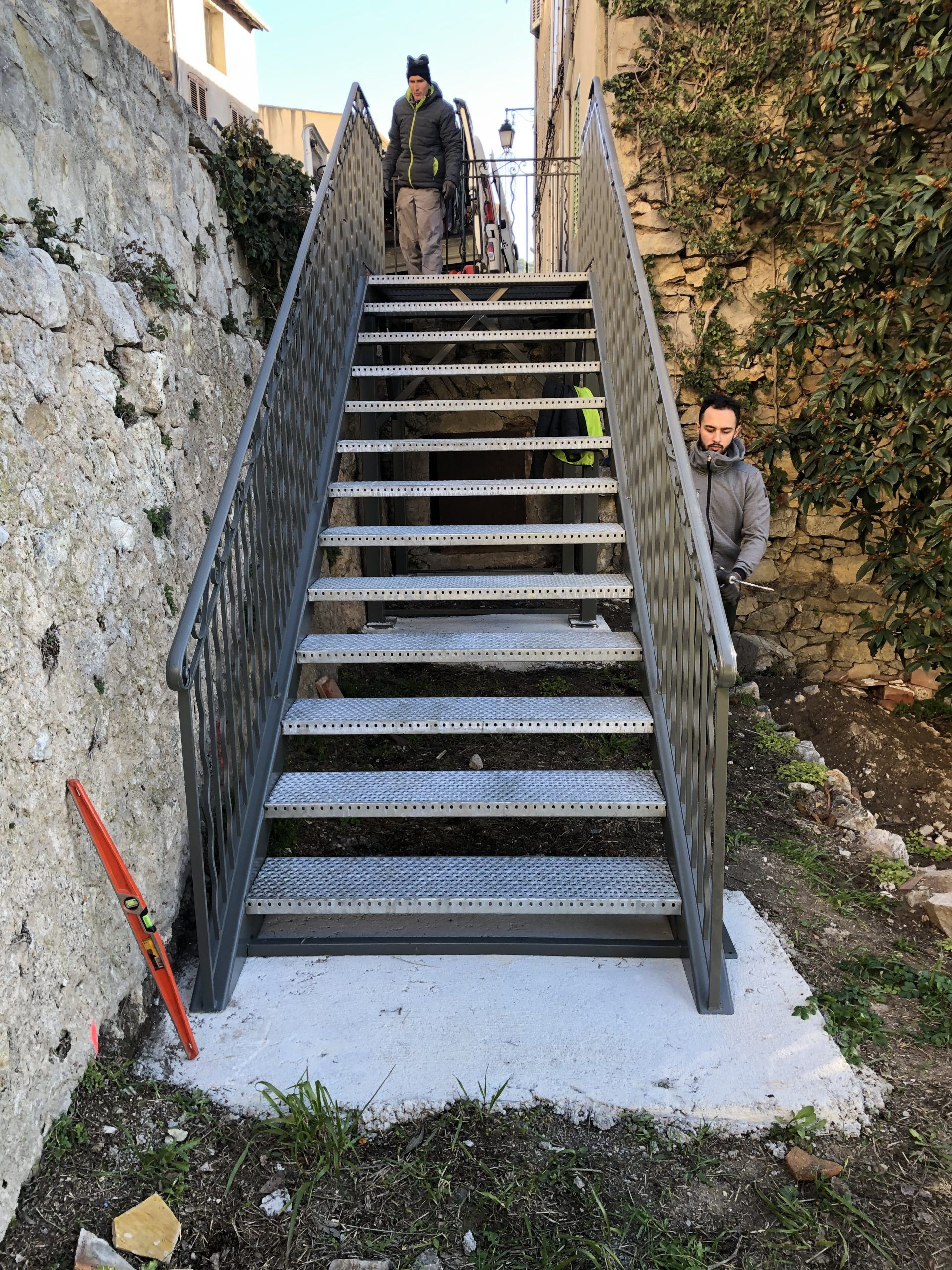 Un escalier métallique abrupt entre des murs de pierre, avec deux personnes debout en haut et une troisième qui regarde sur le côté.