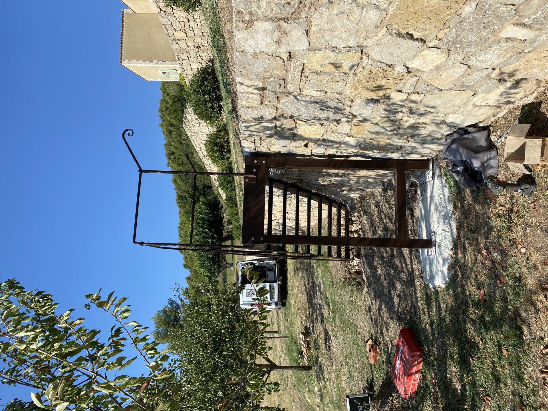 Entrée d'un bâtiment en pierre avec un portail en métal, chemin de terre et un tissu rouge suspendu près d'un jardin sous un ciel bleu.