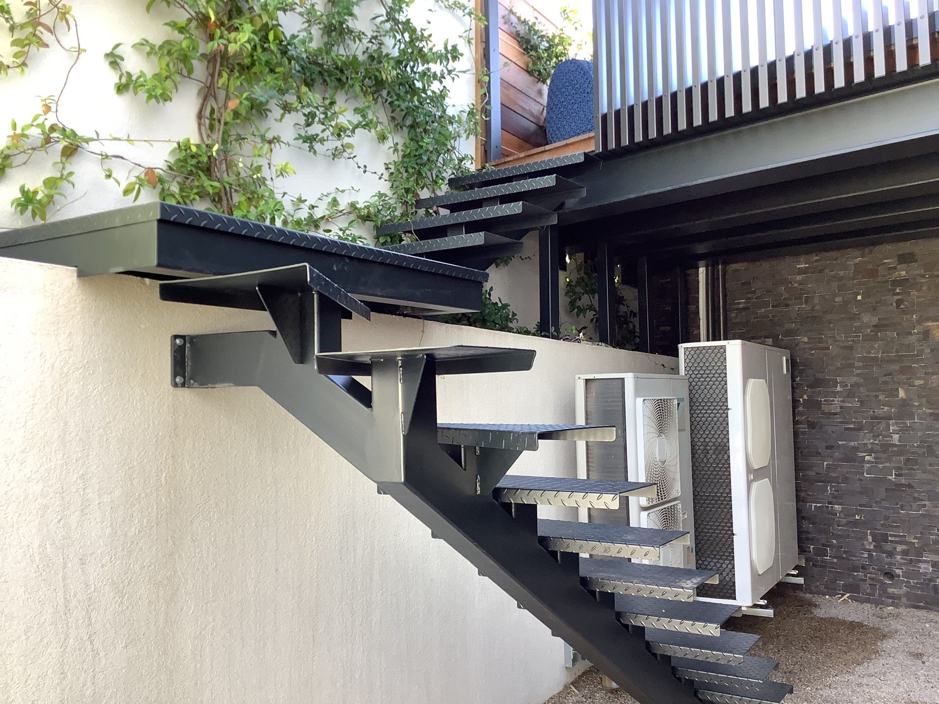 Escalier métallique et passerelle surélevée longeant un mur en stuc beige, avec du lierre et des unités utilitaires stationnées en contrebas.