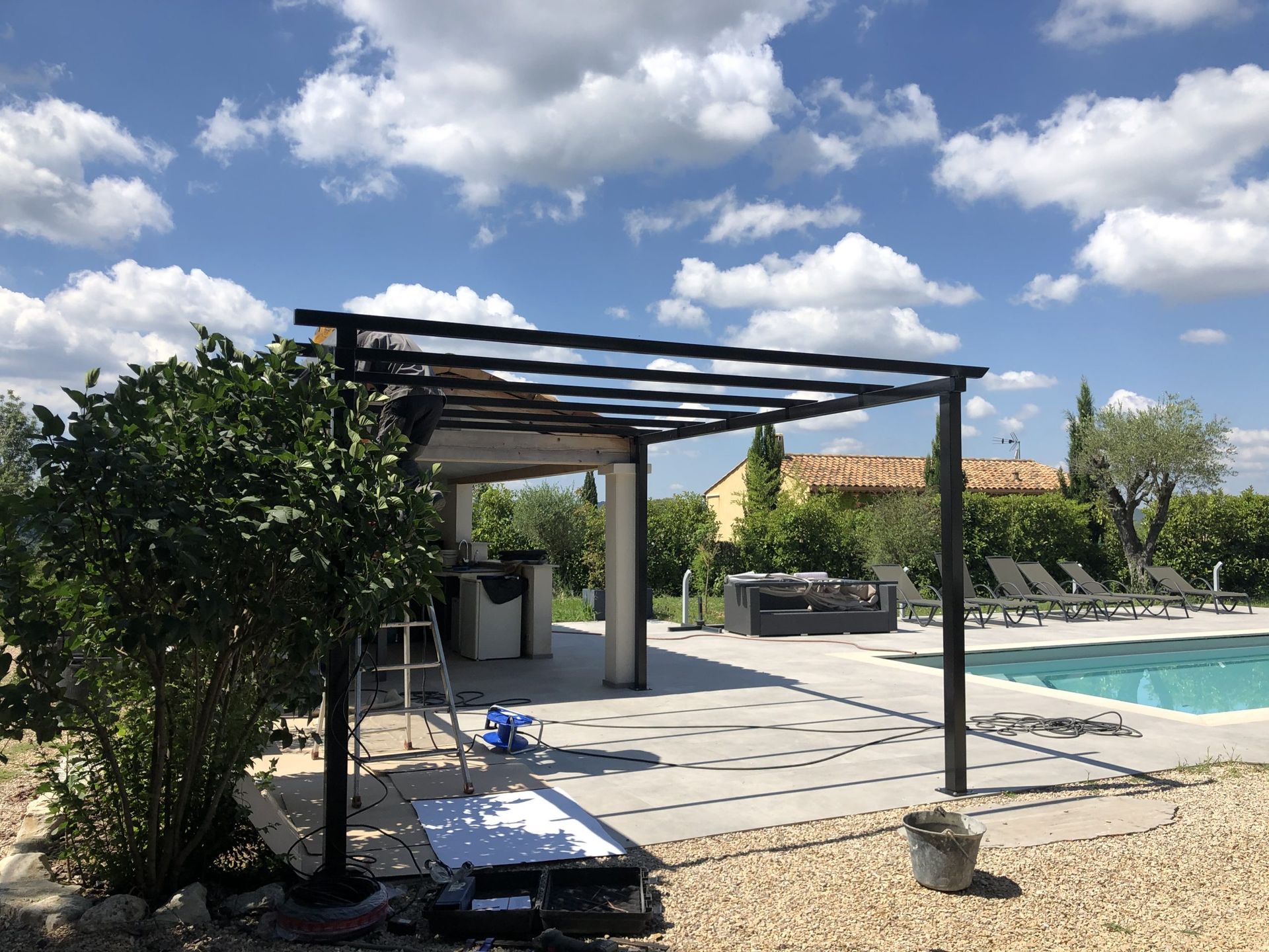 Terrasse au bord de la piscine avec pergola noire, dalles de pierre, verdure et ciel bleu parsemé de nuages