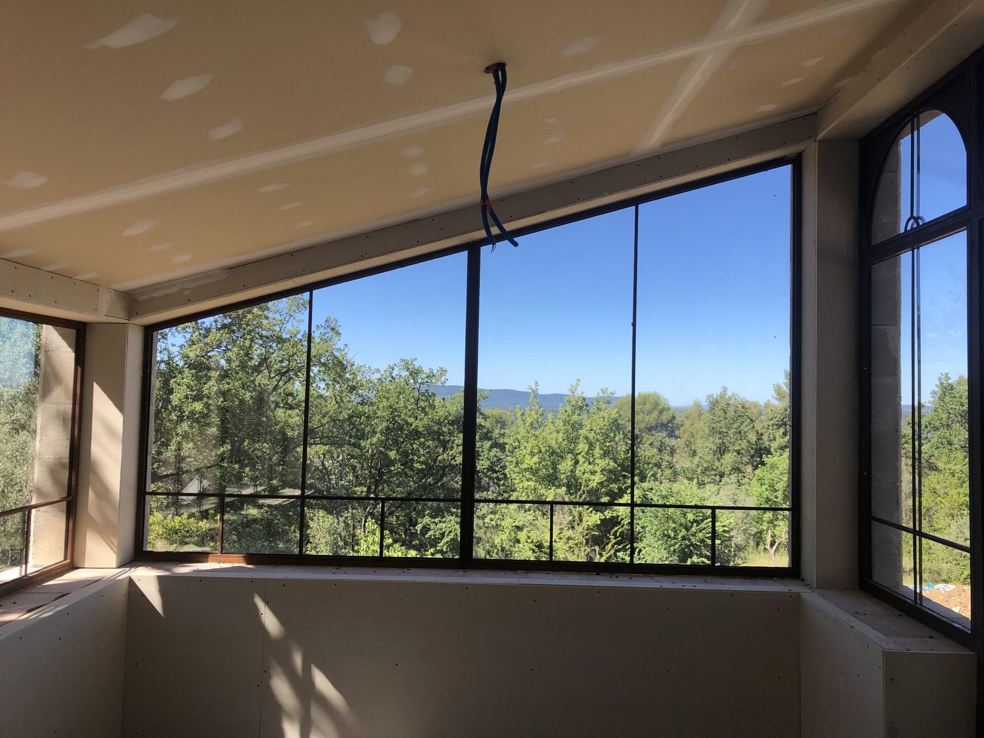 Véranda fermée avec rambarde noire, grandes fenêtres et vue sur des arbres verdoyants et un ciel bleu.
