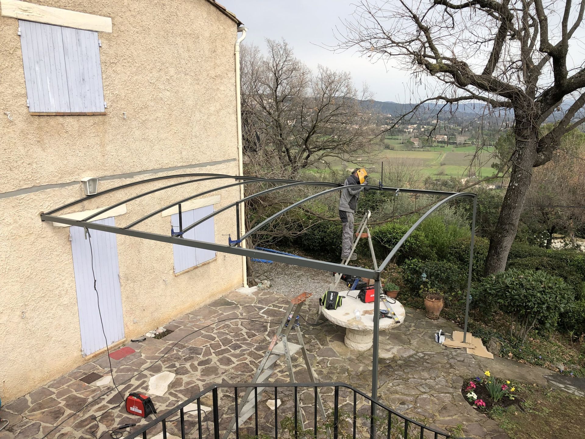 Terrasse arrière attenante à la maison, avec une grande structure métallique en construction au milieu des arbres et de la pelouse.