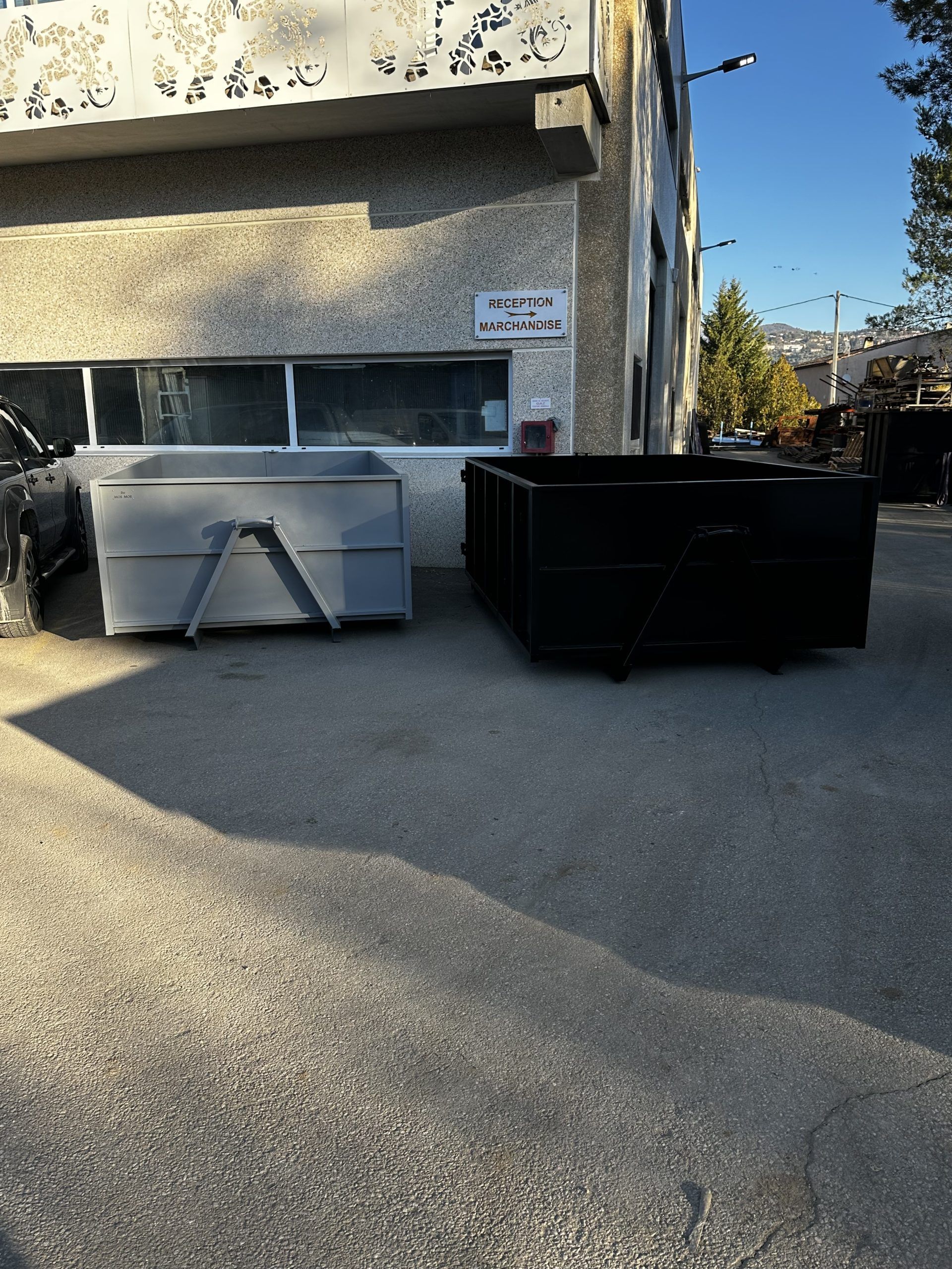 Deux bennes à ordures devant un bâtiment en béton dans une rue ensoleillée