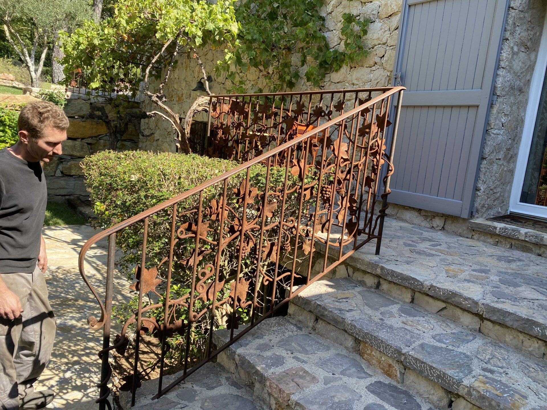 Un homme se tient debout près de marches en pierre et d'une balustrade en fer forgé décorative dans une cour extérieure ensoleillée.