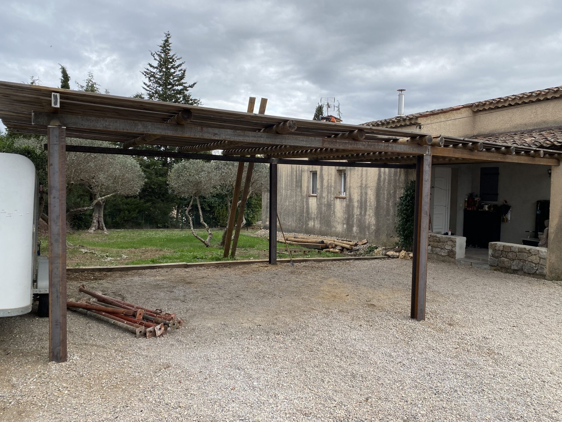 Pergola rustique au-dessus d'une cour de gravier, à côté d'un bâtiment en stuc, sous un ciel nuageux.