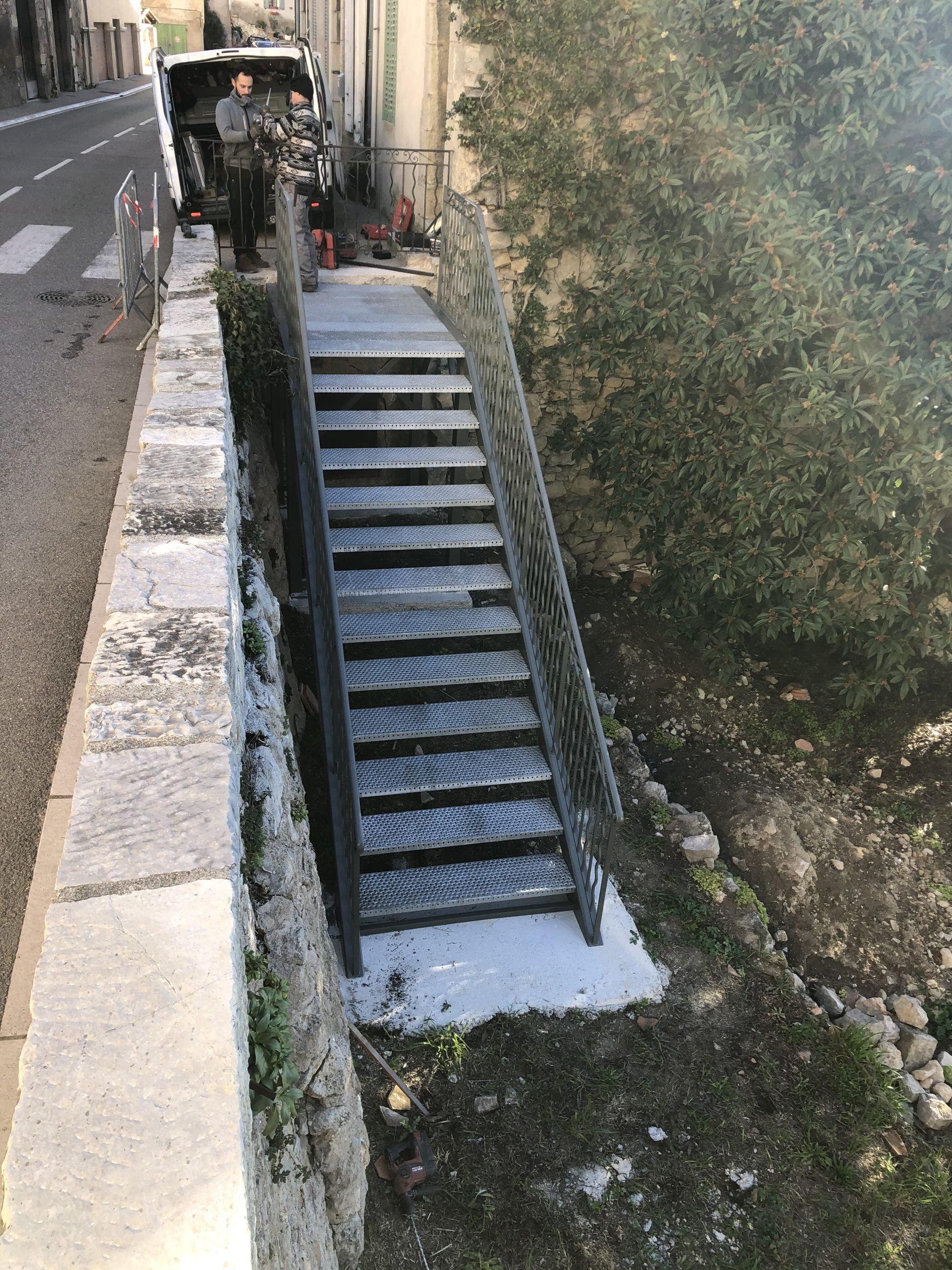 Escalier extérieur étroit descendant le long d'un mur de pierre, avec une rampe et un versant boisé à droite.
