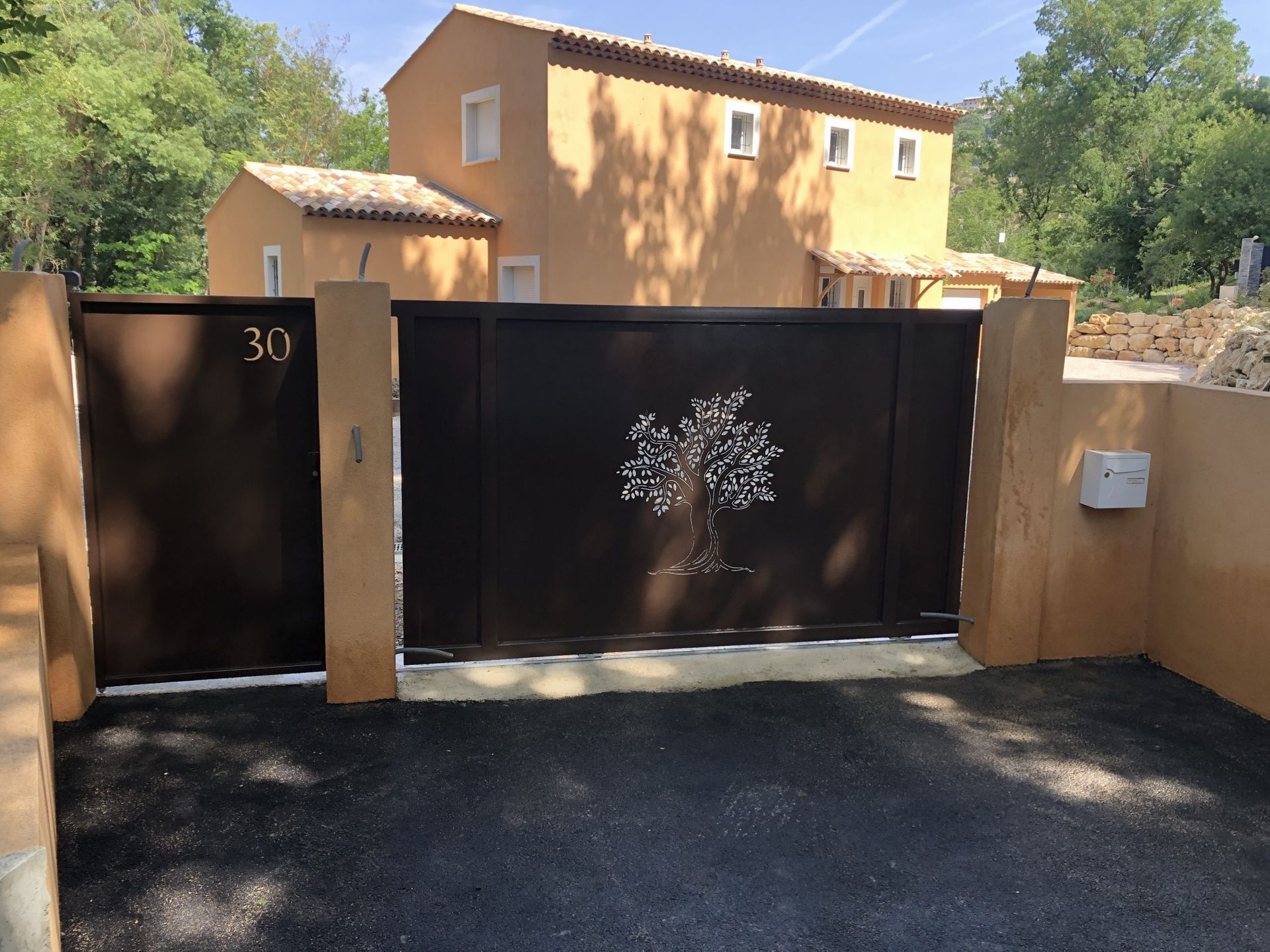 Portail d'allée en métal noir devant une maison beige, avec une boîte aux lettres à droite et des arbres en arrière-plan.