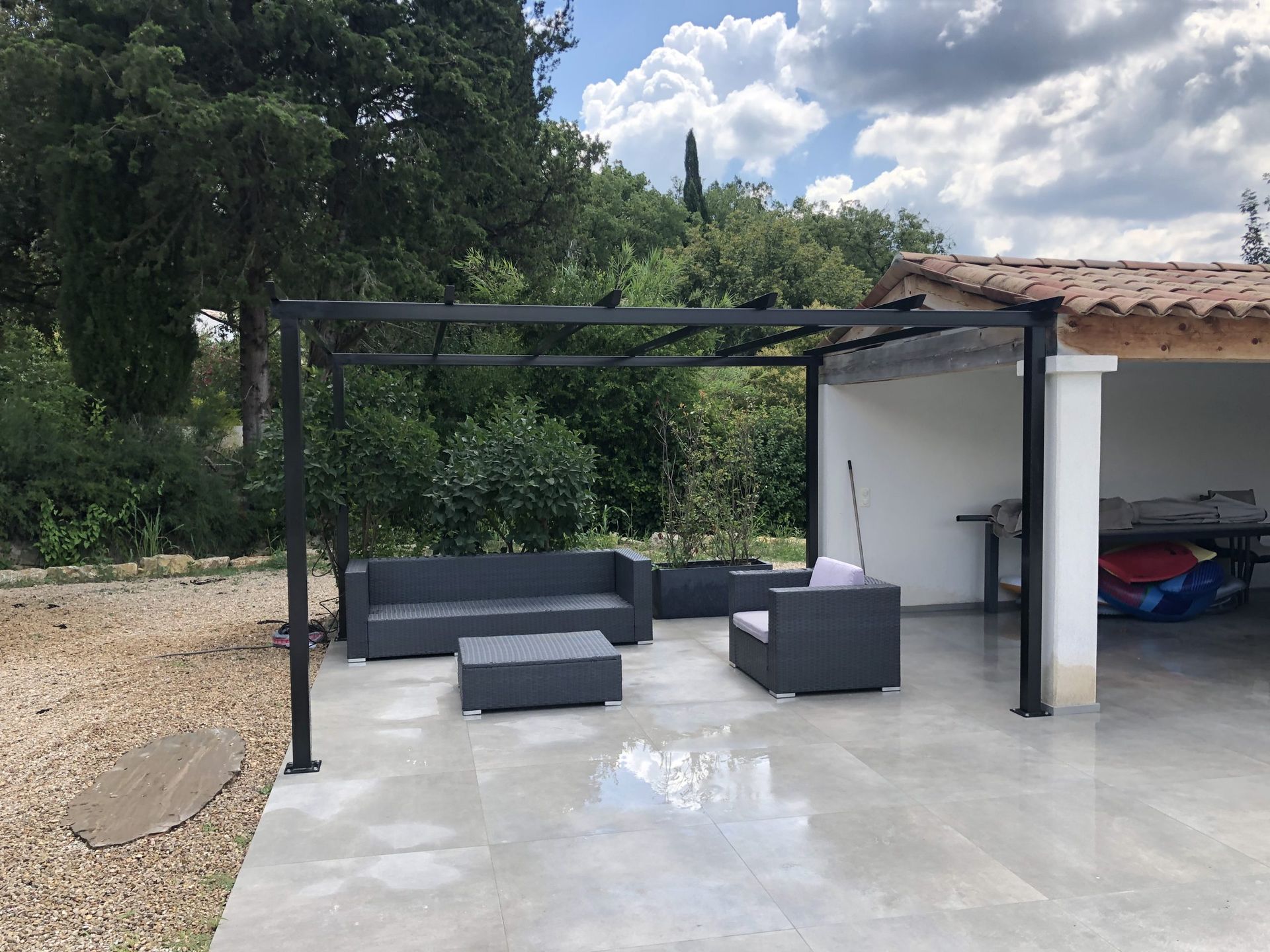 Pergola noire avec sièges d'extérieur gris sur une terrasse en béton à côté d'un abri de voiture, avec des arbres et un ciel bleu en arrière-plan.