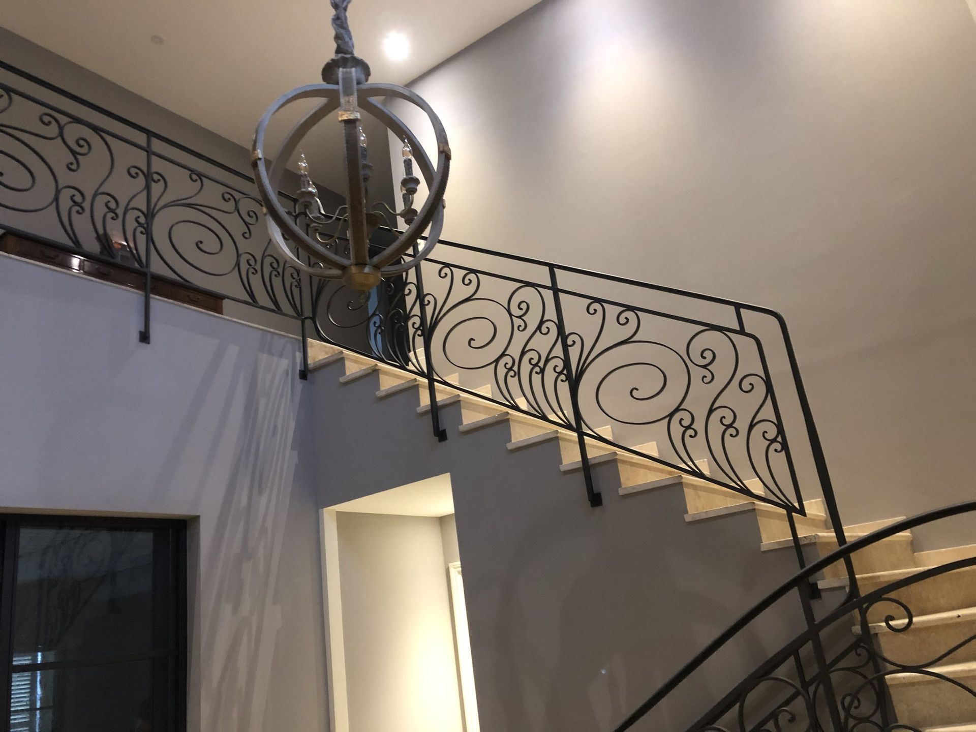 Escalier intérieur avec rampe noire ornée et lustre suspendu sous une lumière chaude