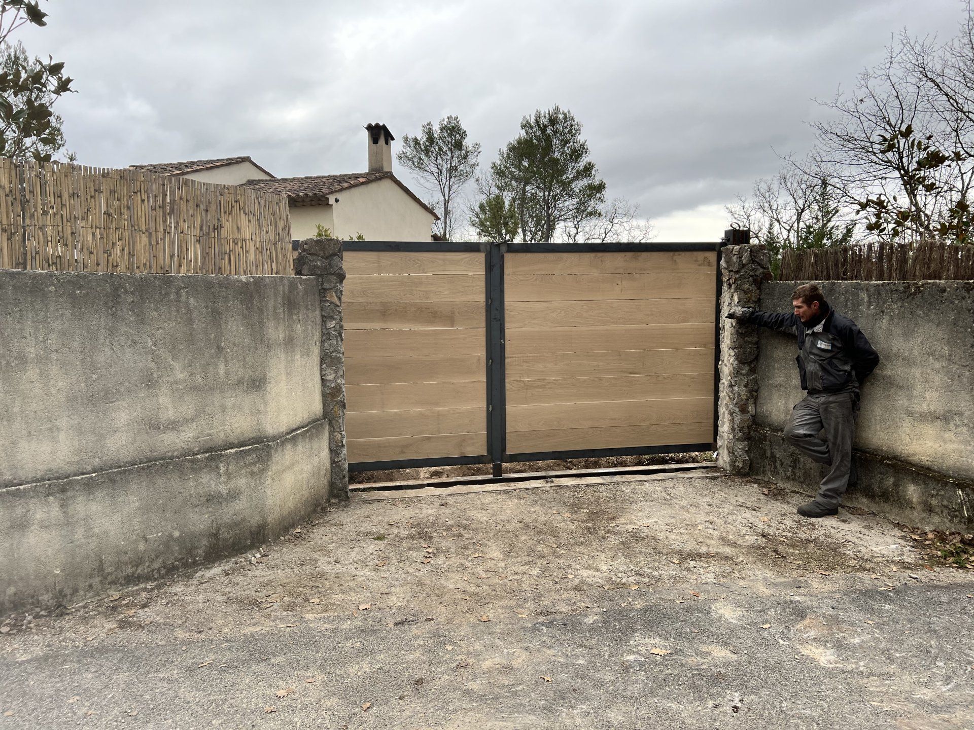Une personne ouvre un portail en bois dans une allée en béton sous un ciel couvert.