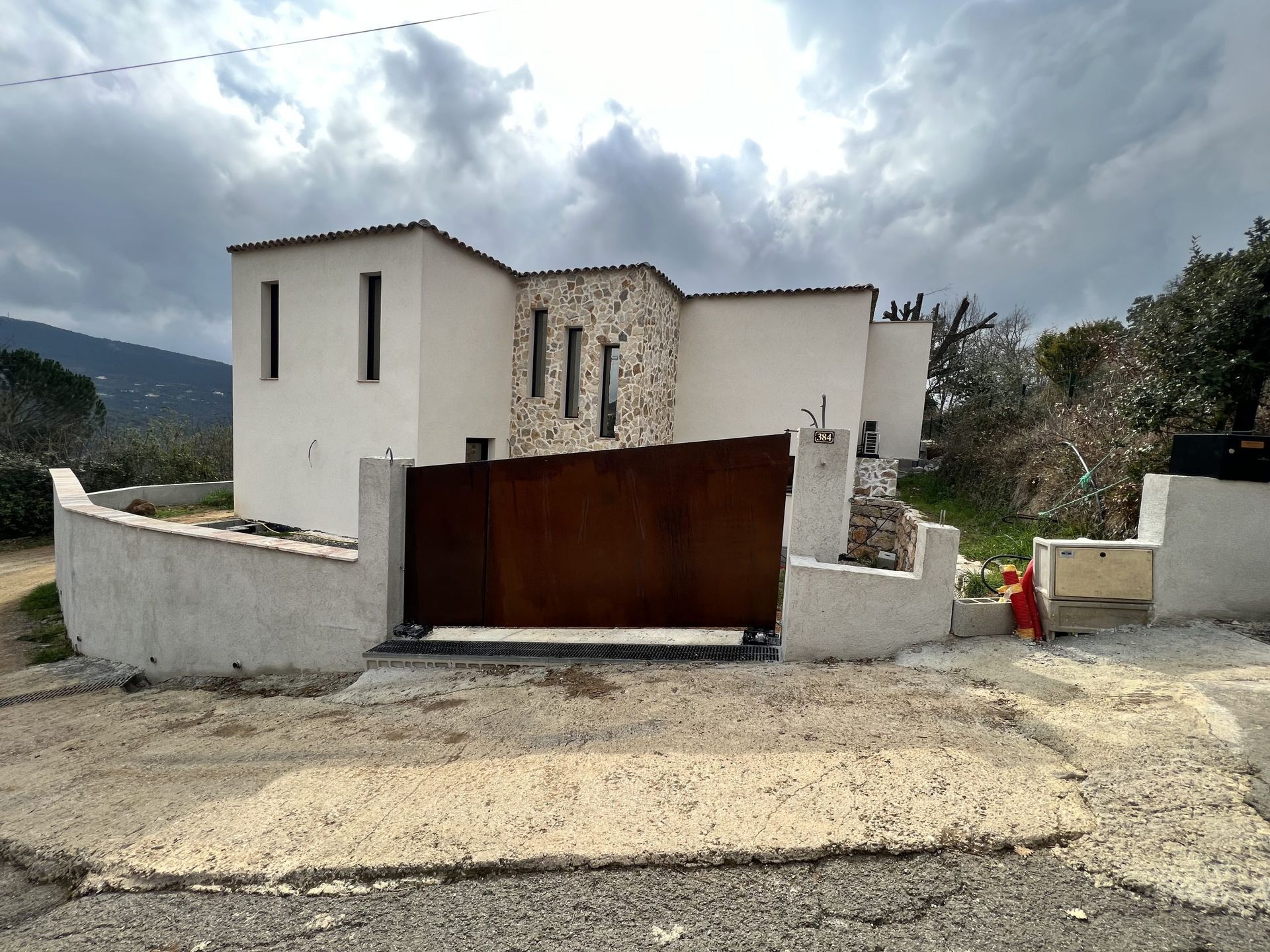 Maison en stuc blanc avec un portail couleur rouille au bord d'un chemin de gravier sous un ciel nuageux.