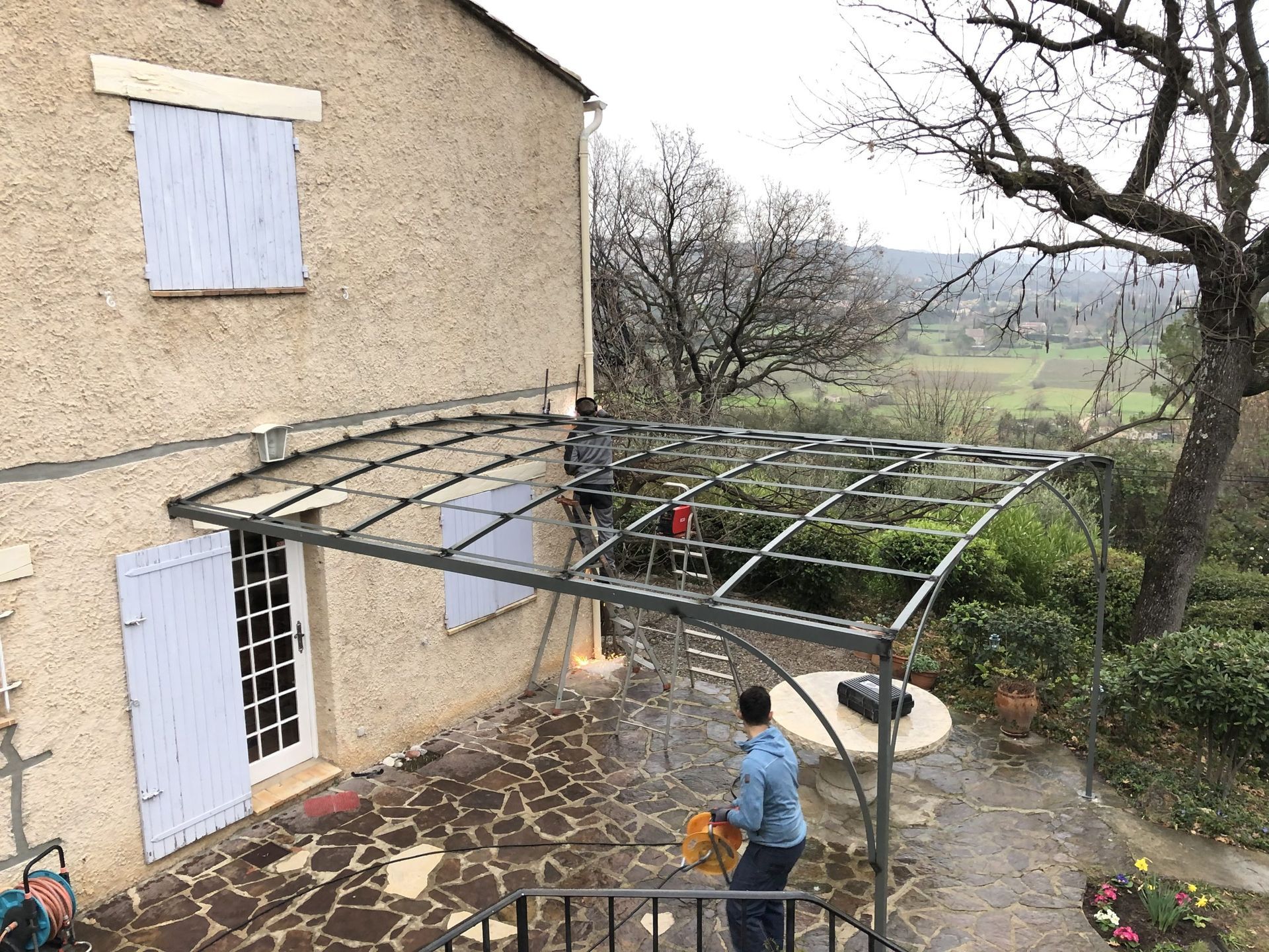 Une personne se trouve sous une pergola métallique à côté d'une maison beige, avec un jardin et des arbres en arrière-plan.