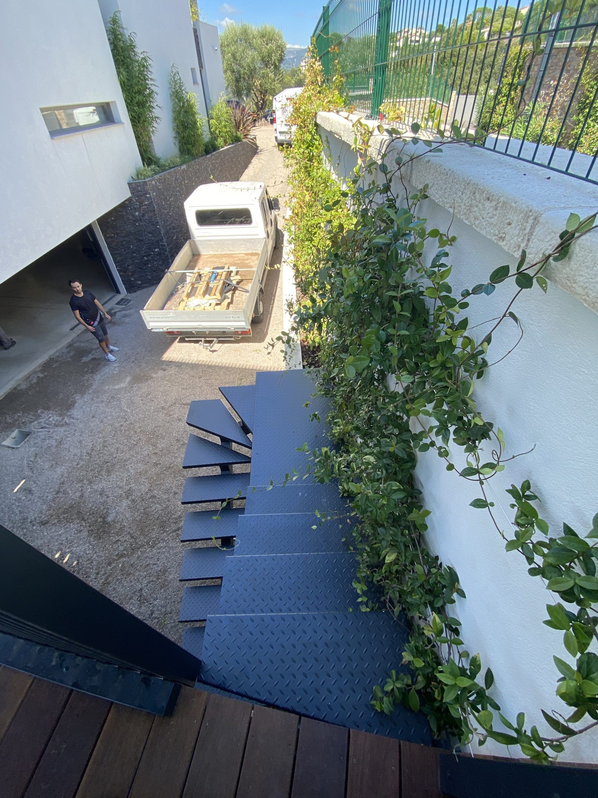 Un étroit escalier extérieur jouxte un bâtiment blanc et mène, en passant devant un camion et de la verdure, à une cour ensoleillée.