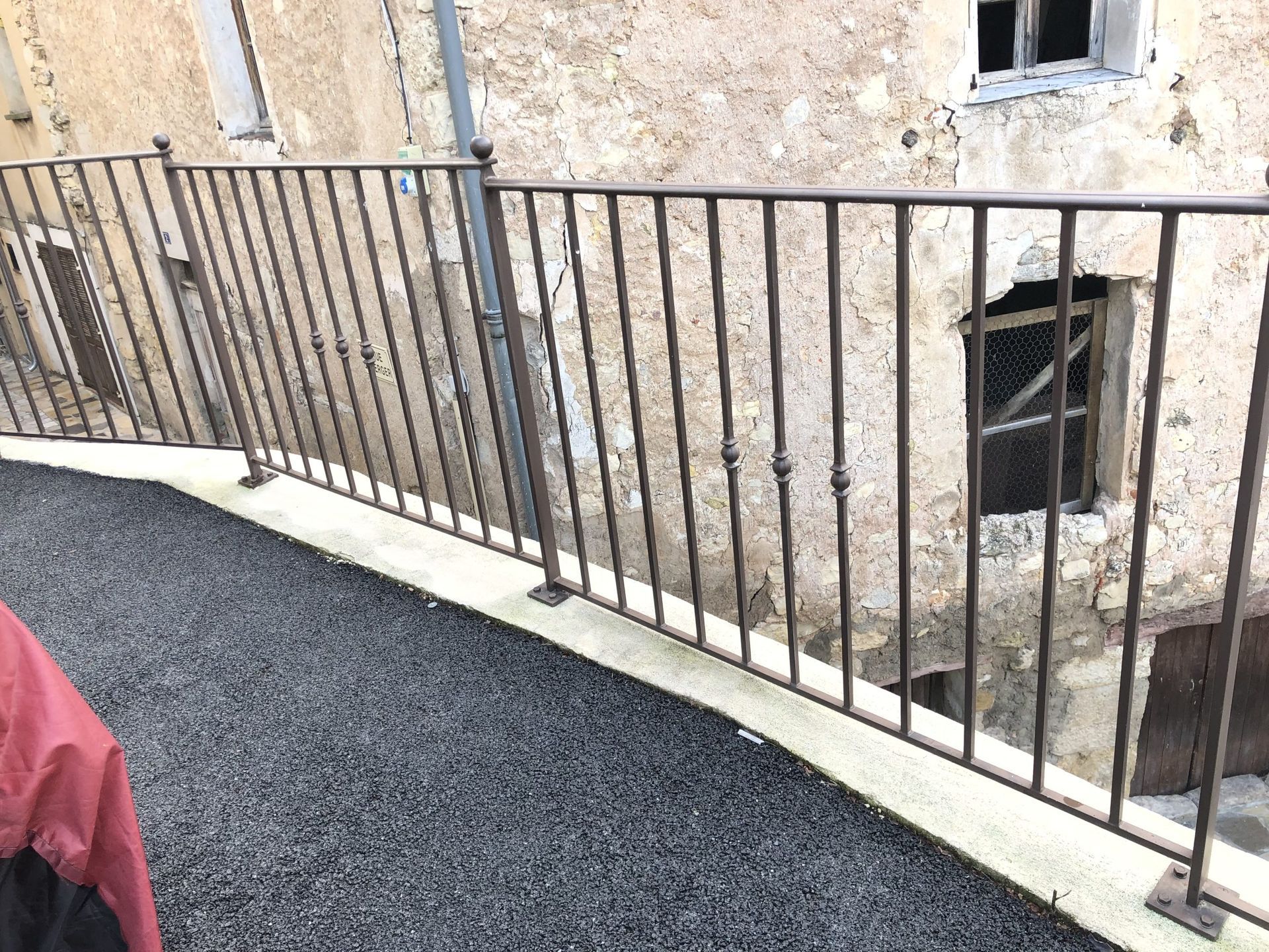 Rambarde en métal noir le long d'un balcon incurvé jouxtant un immeuble en stuc patiné, avec des fenêtres et du gravier en dessous.