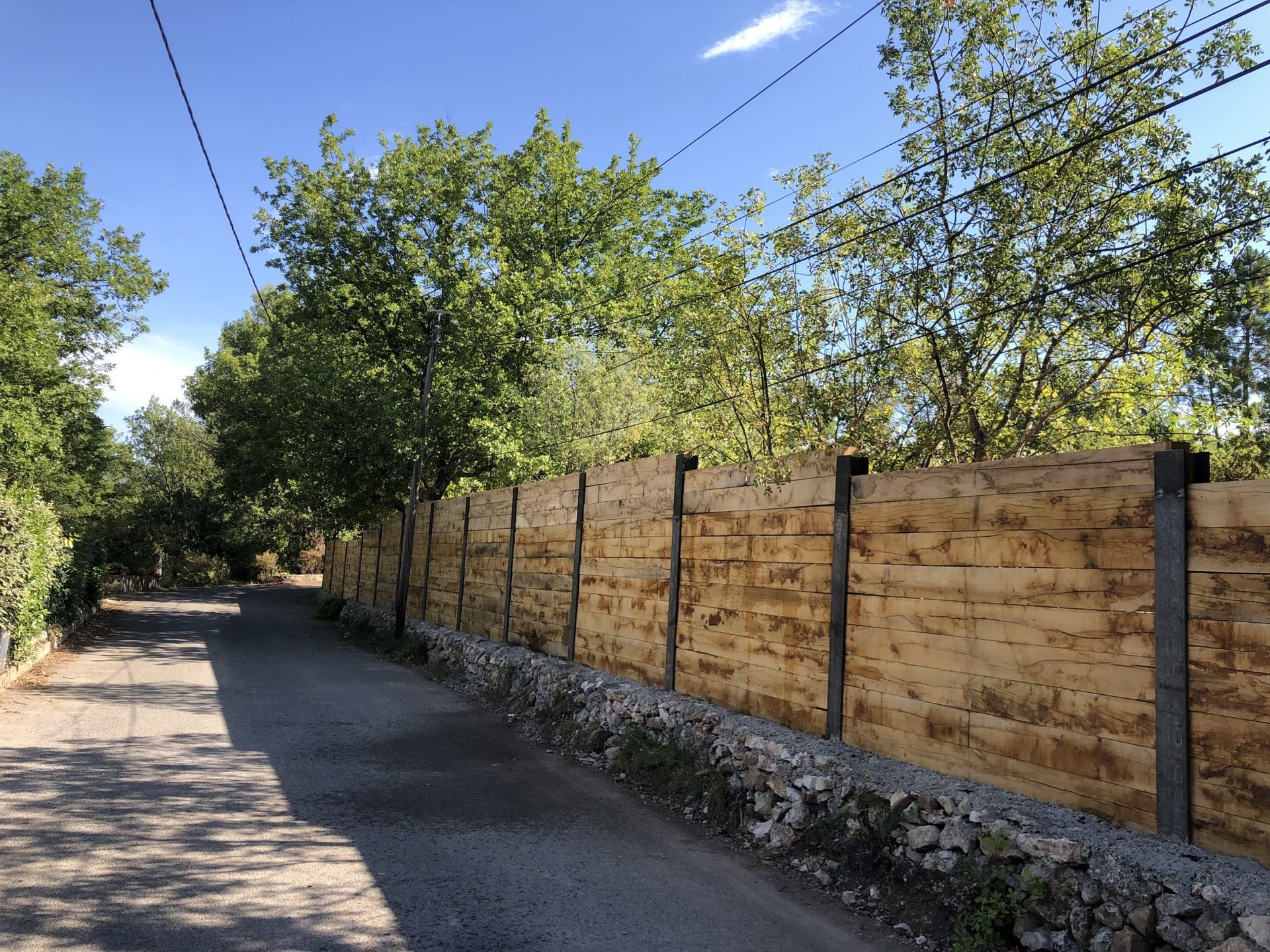Route pavée bordée d'arbres, longeant une clôture en bois sous un ciel bleu
