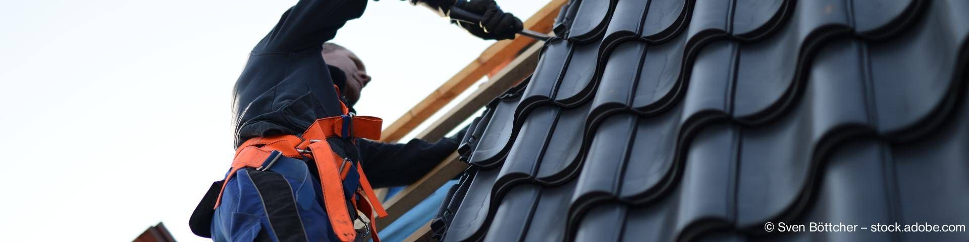 Dachdecker mit Schutzhelm und orangefarbenem Sicherheitsgurt auf schwarzer Dachziegeldeckung