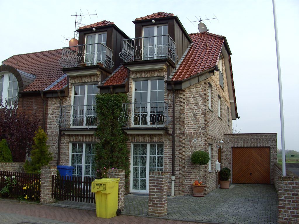Zweistöckiges Backsteinhaus mit roten Dachziegeln, zwei Balkonen, einer Garage und mehreren Mülltonnen vor dem Zaun.