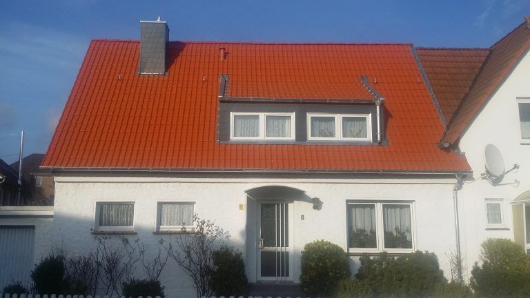 Weißes Einfamilienhaus mit rotem Satteldach, drei Fenstern im Dachgeschoss und einem Eingang mit Glasfront.