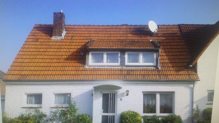 Weißes Einfamilienhaus mit braunem Ziegeldach, Dachgaube mit zwei Fenstern, Schornstein und Satellitenschüssel