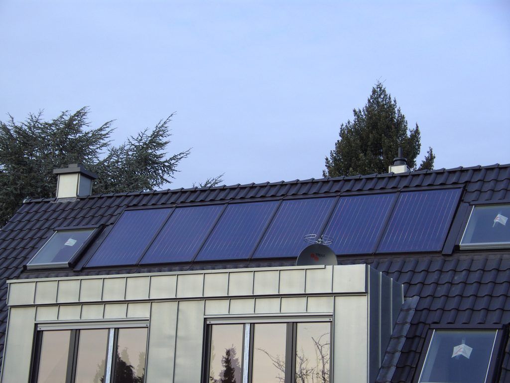 Dach mit sechs rechteckigen Solarpaneelen, zwei Dachfenstern und Satellitenschüssel, dahinter Bäume und blauer Himmel.