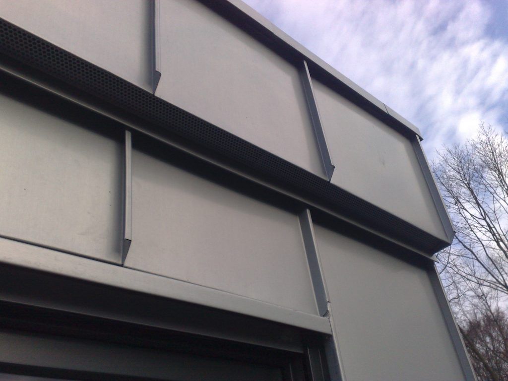 Nahaufnahme einer grauen Metallfassade mit vertikalen Verstrebungen, im Hintergrund blauer Himmel und Baumzweige.