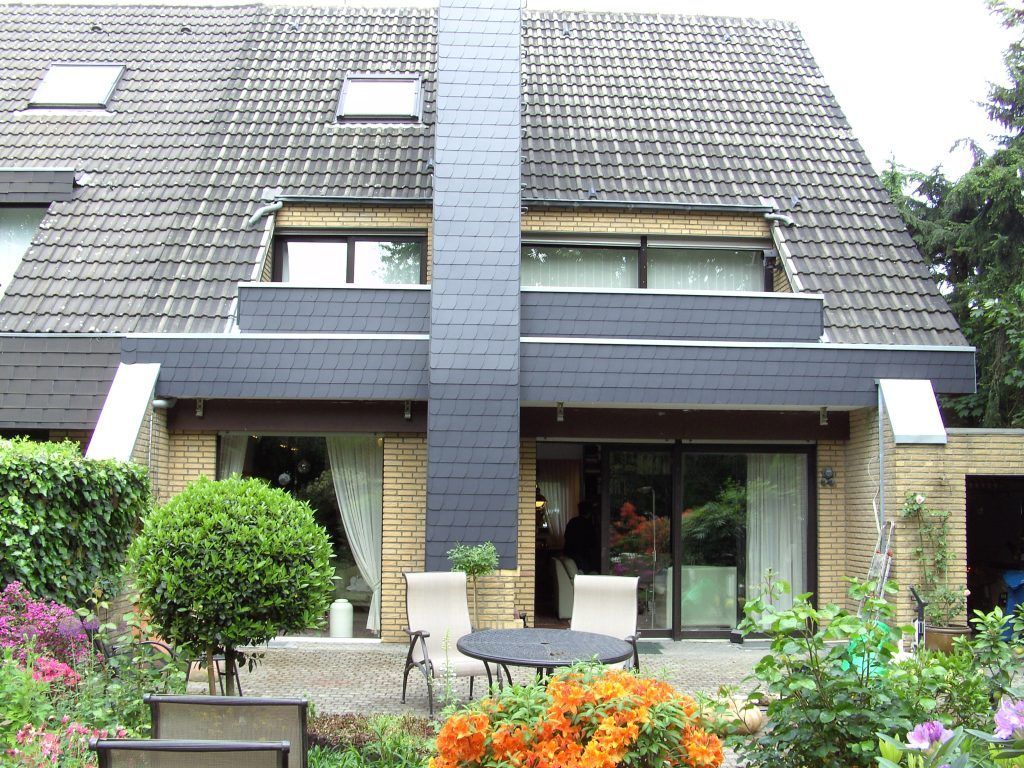 Zweistöckiges Haus mit Satteldach, Terrasse mit Gartentisch und Stühlen, umgeben von Blumen und Sträuchern.