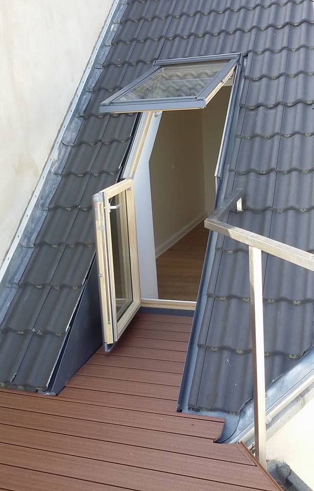 Geöffnetes Dachfenster mit Klapp- und Schiebeteil in einem Dach mit dunklen Ziegeln, davor Holzterrasse mit Glasgeländer.