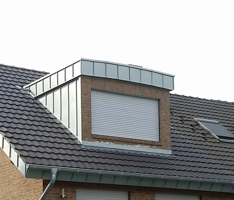 Dachgaube mit geschlossener weißer Rollladenklappe, Ziegelfassade und dunklen Dachziegeln, daneben Dachfenster.
