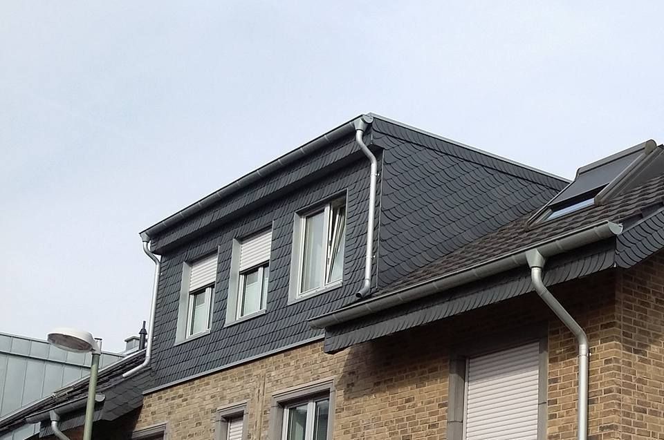 Dachgaube mit Schieferverkleidung und drei Fenstern auf einem Backsteinhaus unter bewölktem Himmel.