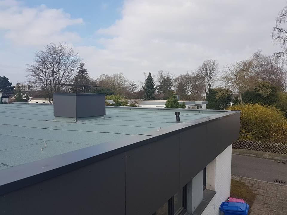 Flachdach mit grauer Dachhaut und kleinem Aufsatz, im Hintergrund Bäume und Wohnhäuser unter bewölktem Himmel.