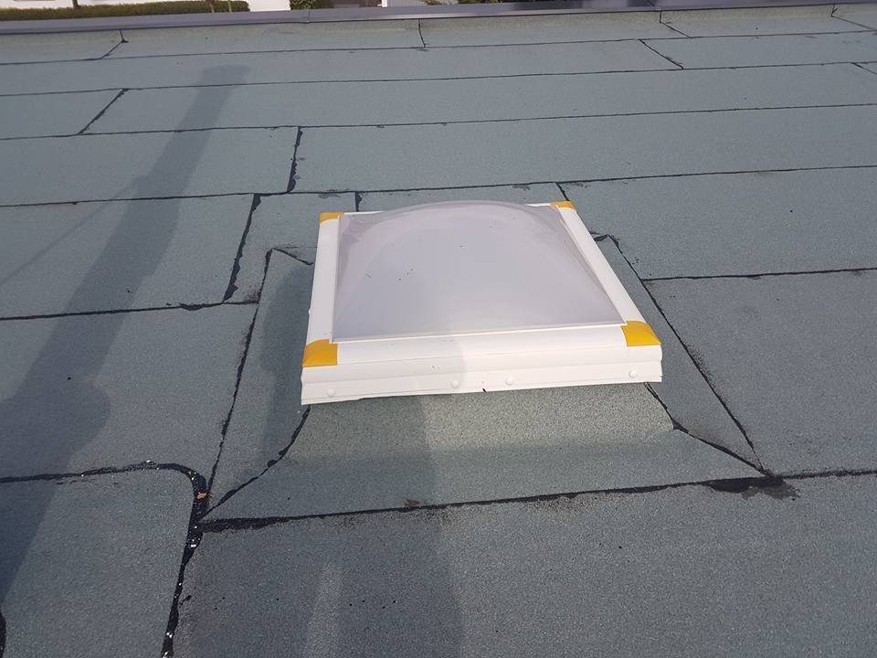 Flaches Dach mit grauer Bitumenabdichtung und quadratischem Dachfenster mit weißem Rahmen und gelben Eckstücken.