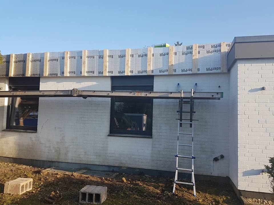 Hauswand mit zwei Fenstern, darauf eine Baustelle mit Dämmplatten und einer Leiter, die an der Wand lehnt.