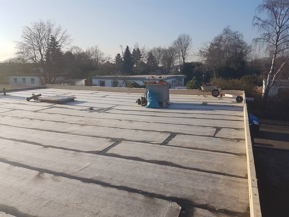 Flachdach mit grauer Abdichtung und schwarzen Nahtstreifen, im Hintergrund Bäume und Wohnhäuser unter klarem Himmel.