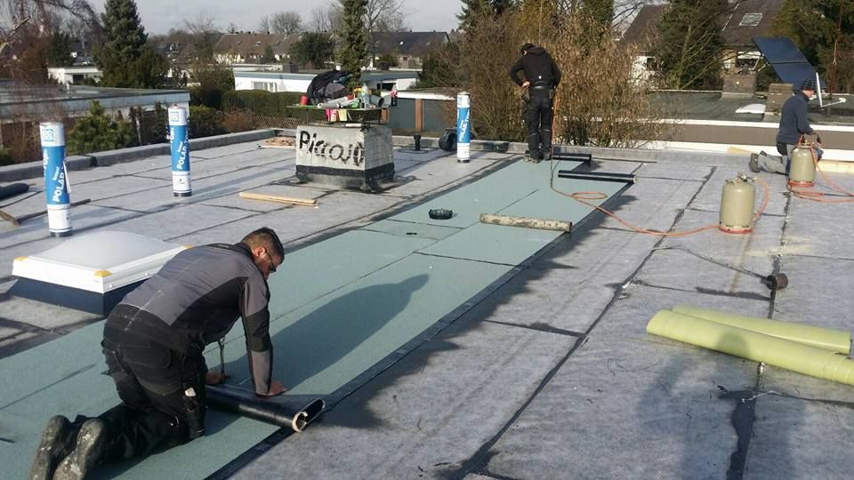 Dachdecker verlegen Dachbahn auf Flachdach, umgeben von Werkzeug und Gasflaschen, bei Tageslicht im Wohngebiet.