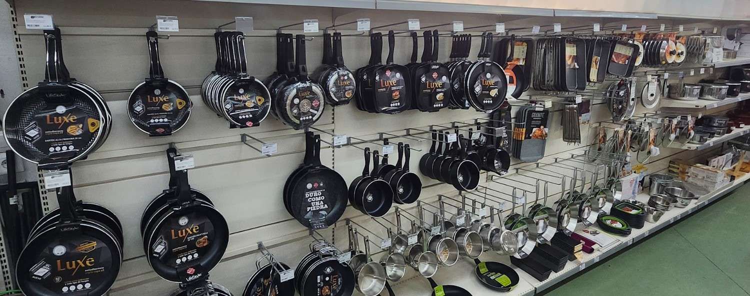 Pared repleta de sartenes y utensilios de cocina colgados en un pasillo de una tienda, con más ollas y utensilios debajo.