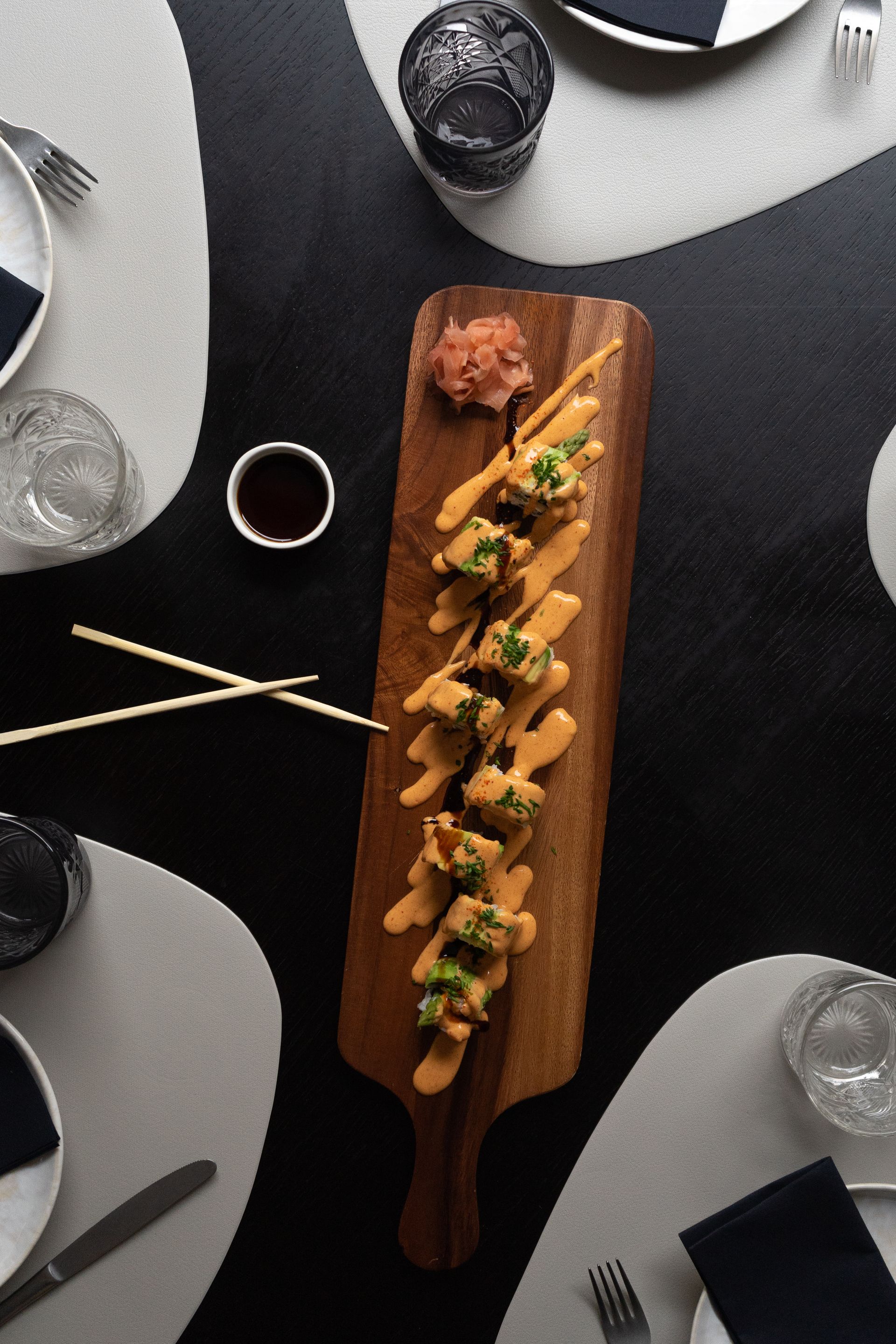 Una larga tabla de cortar de madera llena de comida y palillos sobre una mesa.