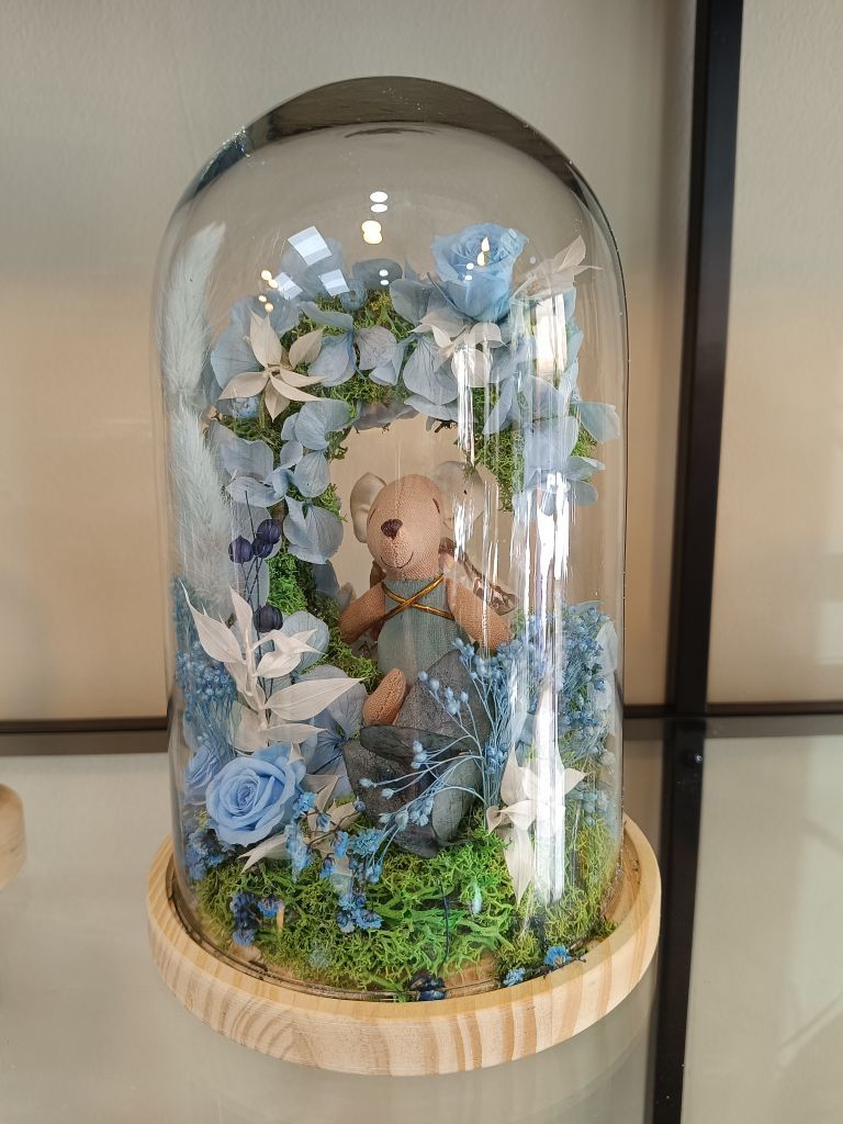 Osito de peluche rodeado de flores azules dentro de una cúpula de cristal sobre una base de madera.