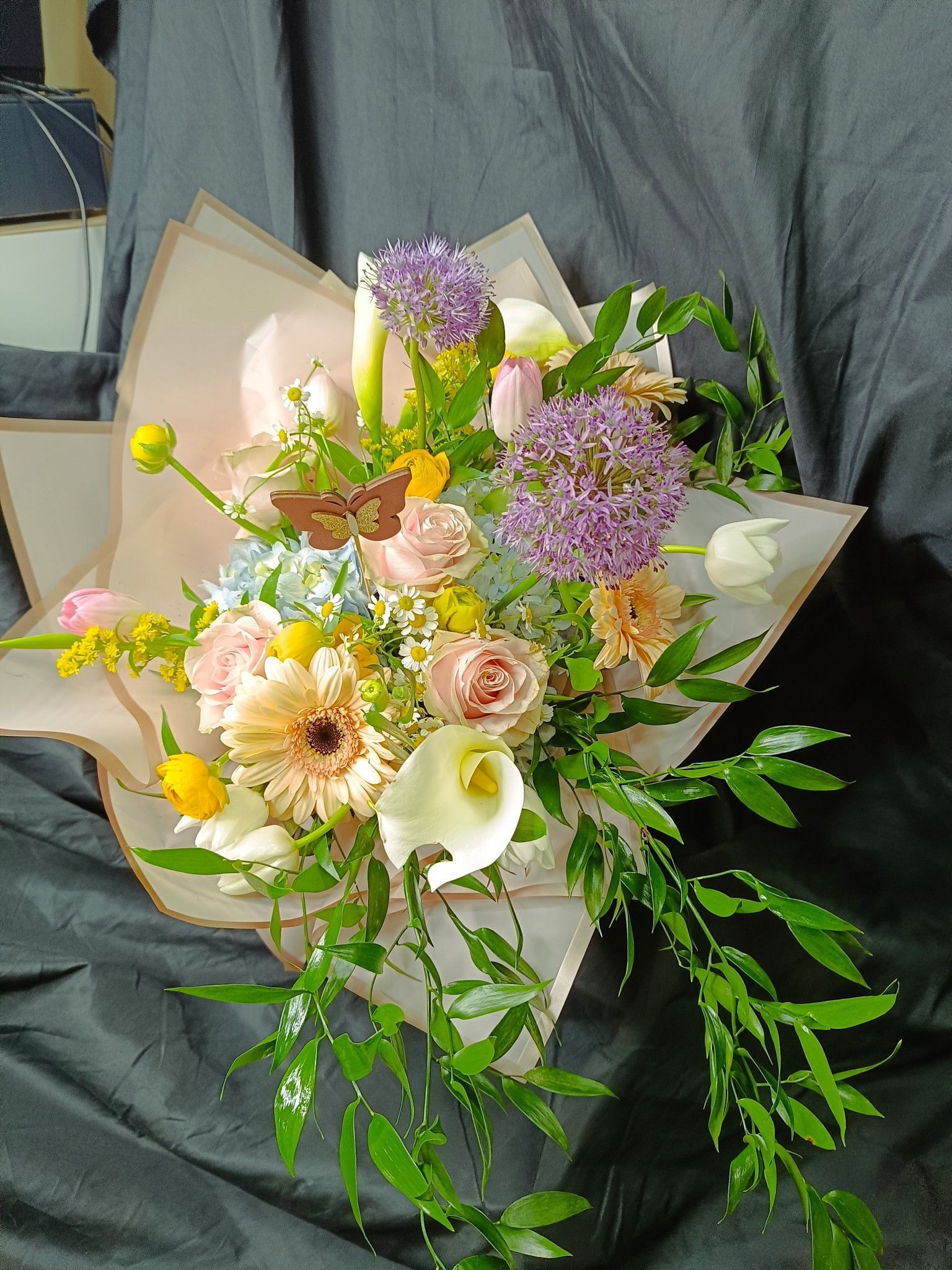 Ramo de flores de colores que incluye rosas, calas y alliums, envuelto en papel, sobre un fondo oscuro.