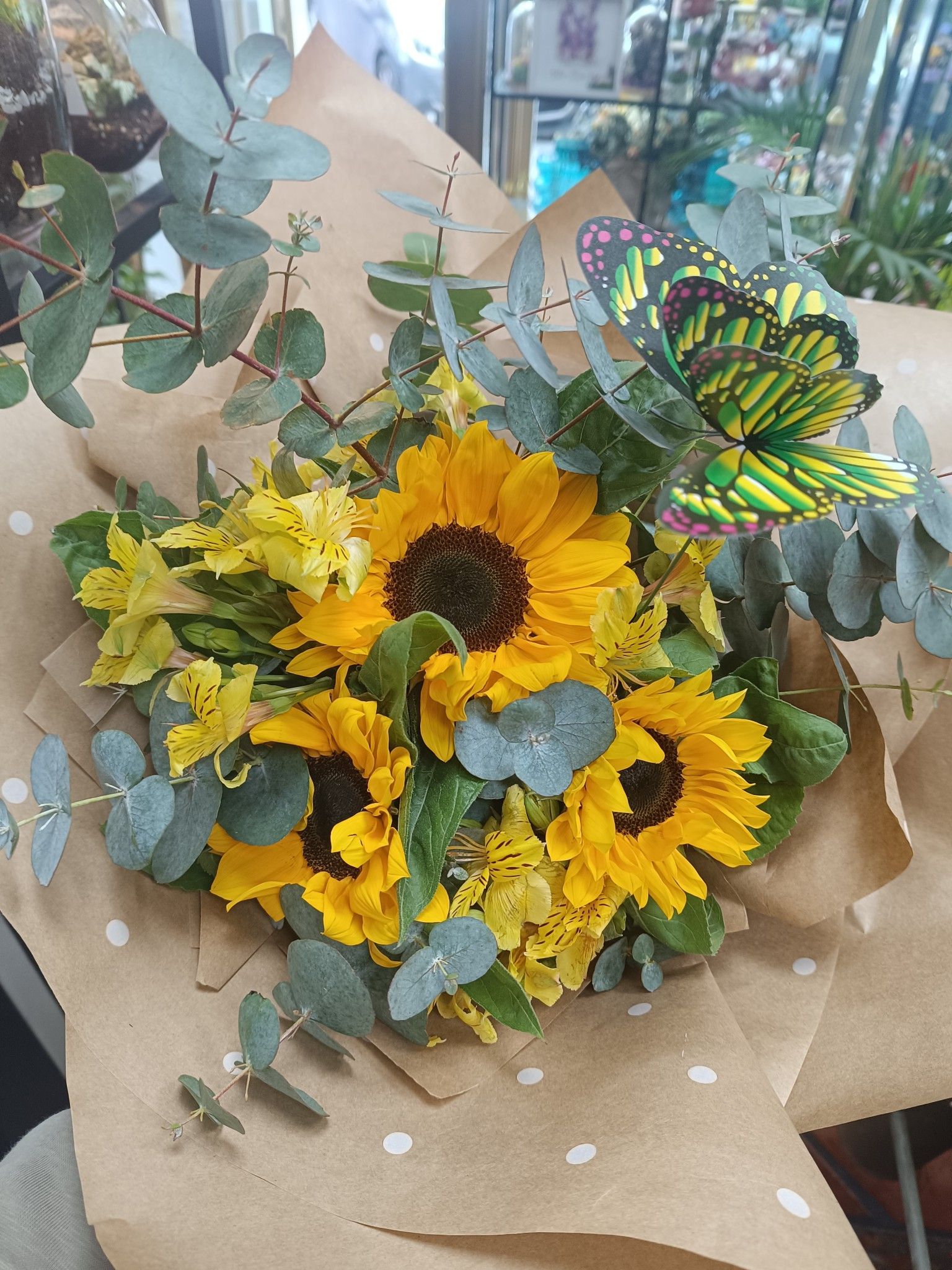 Ramo de girasoles y flores amarillas envuelto en papel marrón, con eucalipto y una mariposa decorativa.