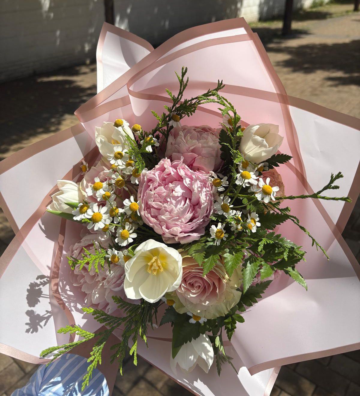 Ramo de flores rosas y blancas envuelto en papel rosa, realizado al aire libre.