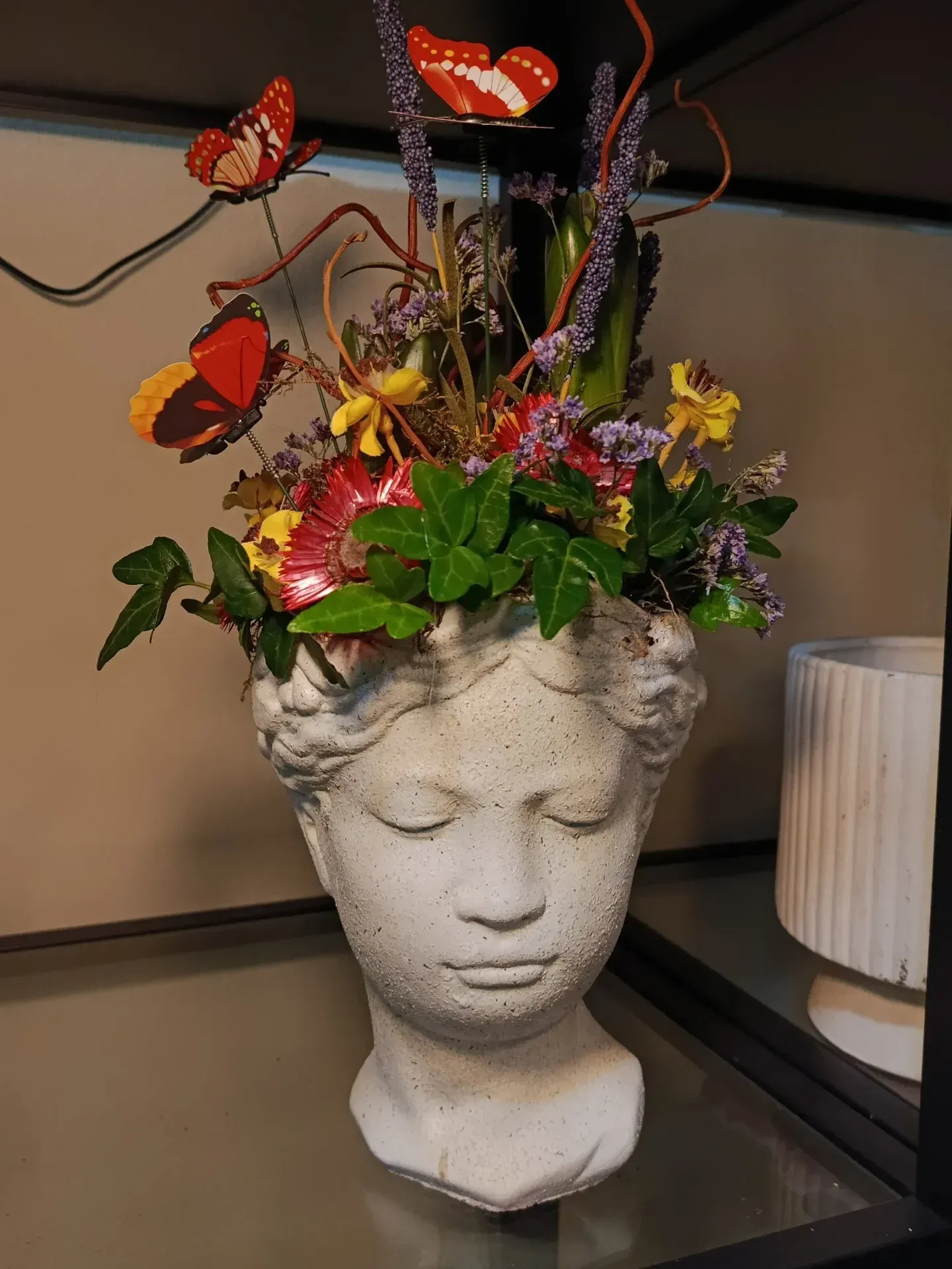 Maceta decorativa blanca con forma de busto, adornada con coloridas flores y mariposas sobre un estante.