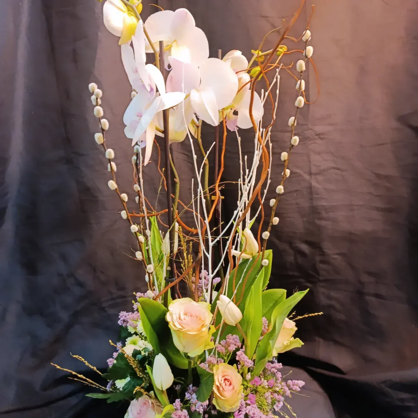 Arreglo floral de orquídeas blancas y rosas rosadas con hojas verdes sobre un fondo oscuro drapeado.