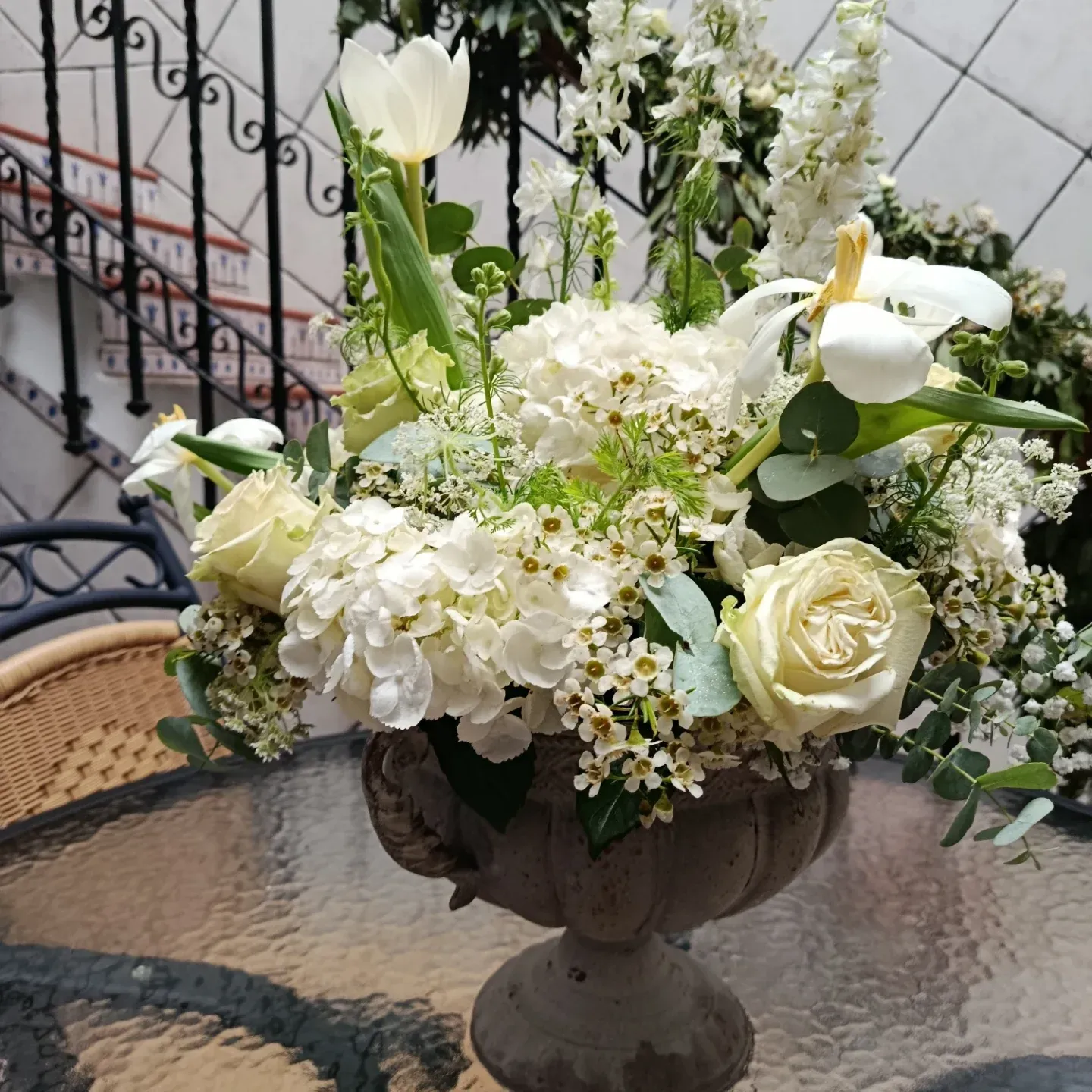 Arreglo floral blanco en una urna de piedra sobre una mesa de patio de cristal con una barandilla de hierro forjado detrás.